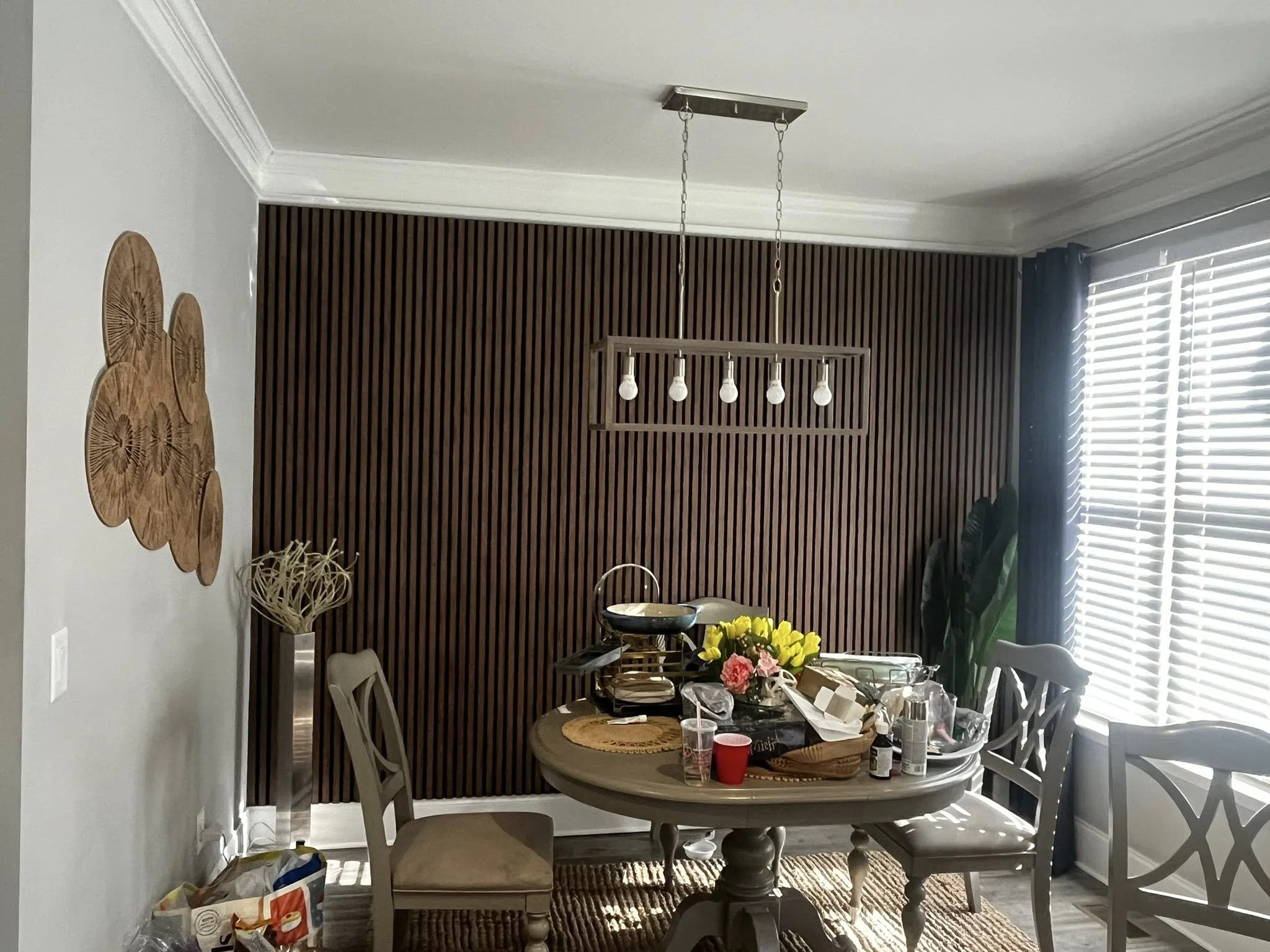 A dining room with a wooden slat accent wall, a round dining table cluttered with various items, chairs around the table, window blinds on the right, and decorative wall art on the left.