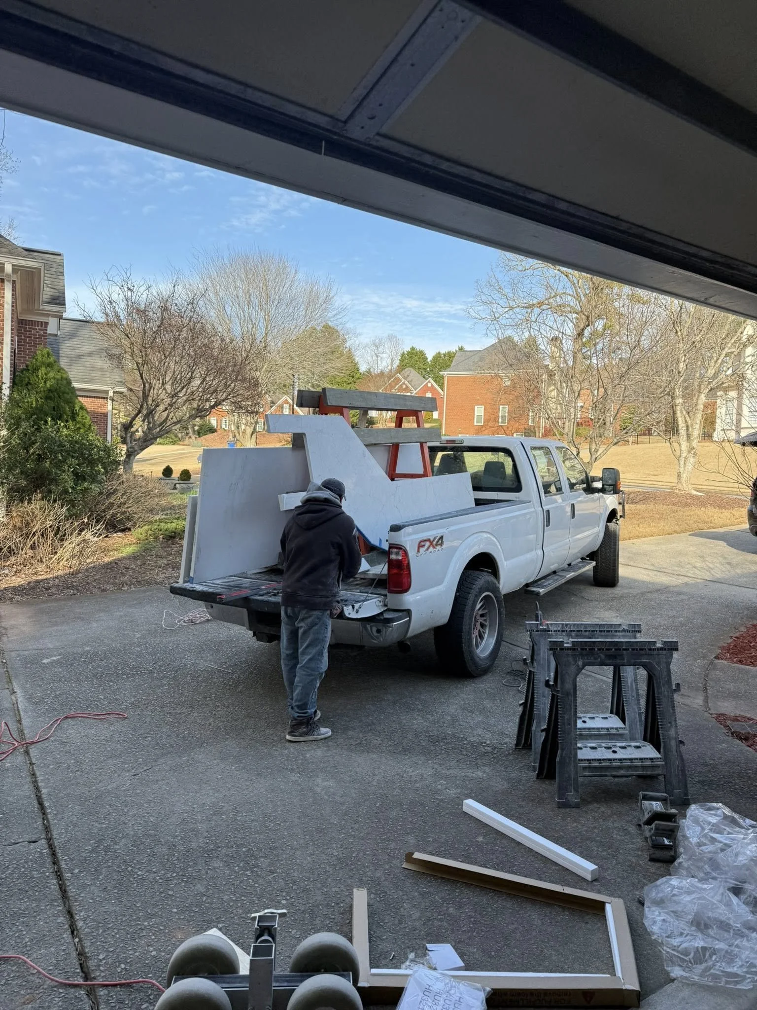 Person working on a white pickup truck parked in a driveway, with construction tools and materials nearby, in a suburban neighborhood with houses and trees.
