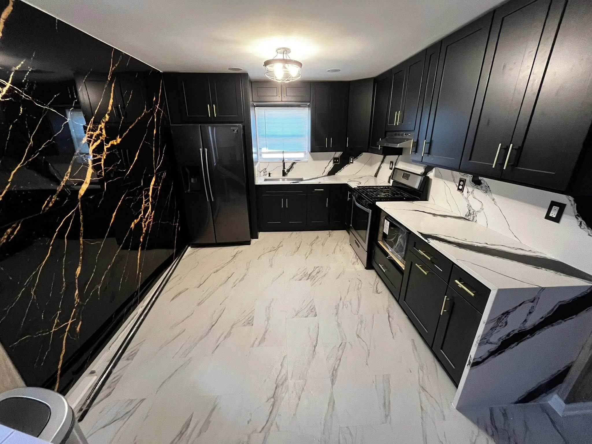 Modern kitchen with black cabinets, white marble countertops with gray and black veining, and black appliances including refrigerator and oven. Black marble accent wall and beige marble flooring.