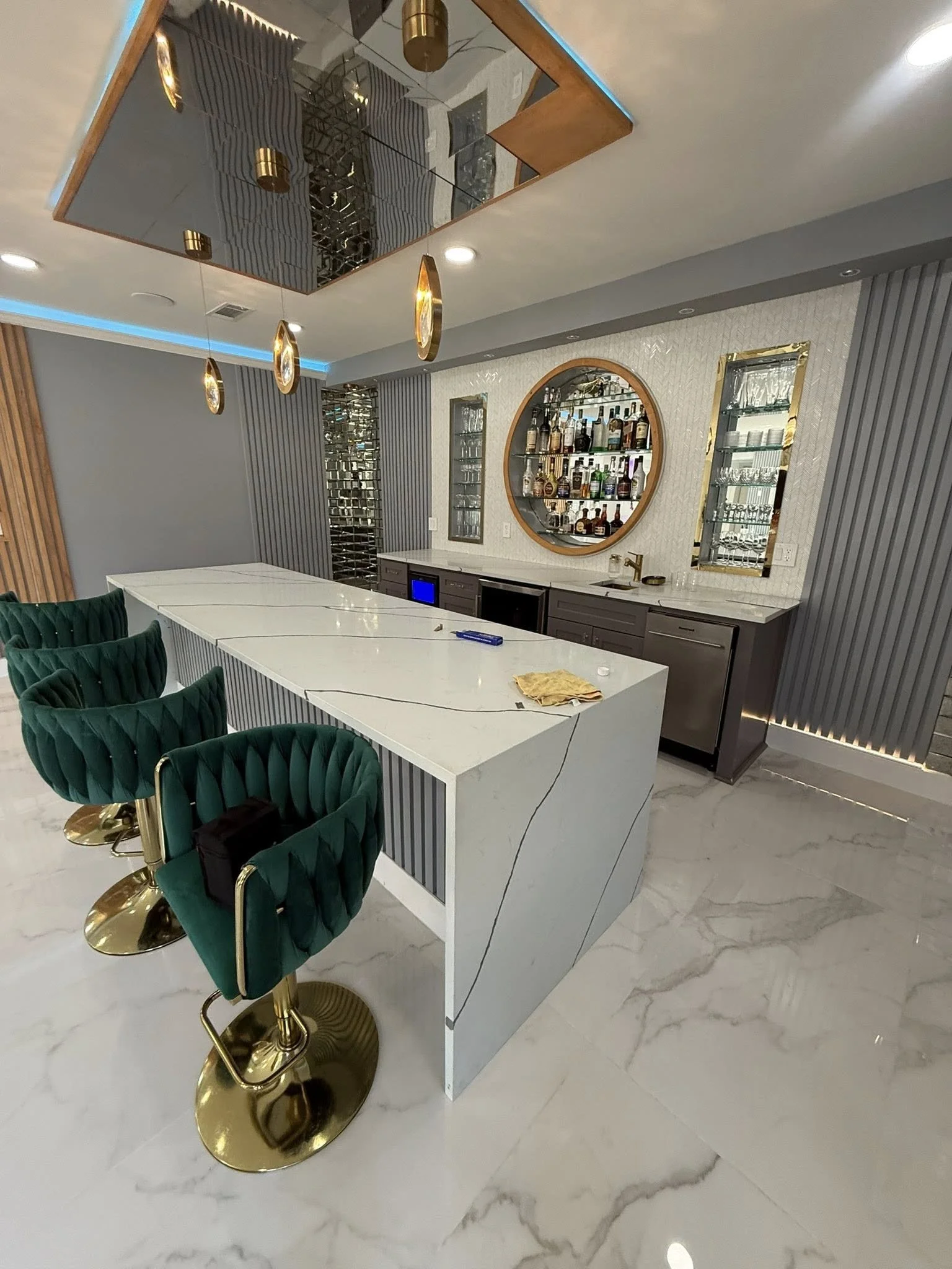Modern home bar area with a white marble countertop, four green velvet bar stools with gold bases, and a wall-mounted circular mirror displaying various bottles of liquor, with two narrow vertical mirrors beside it. The ceiling has a large reflective