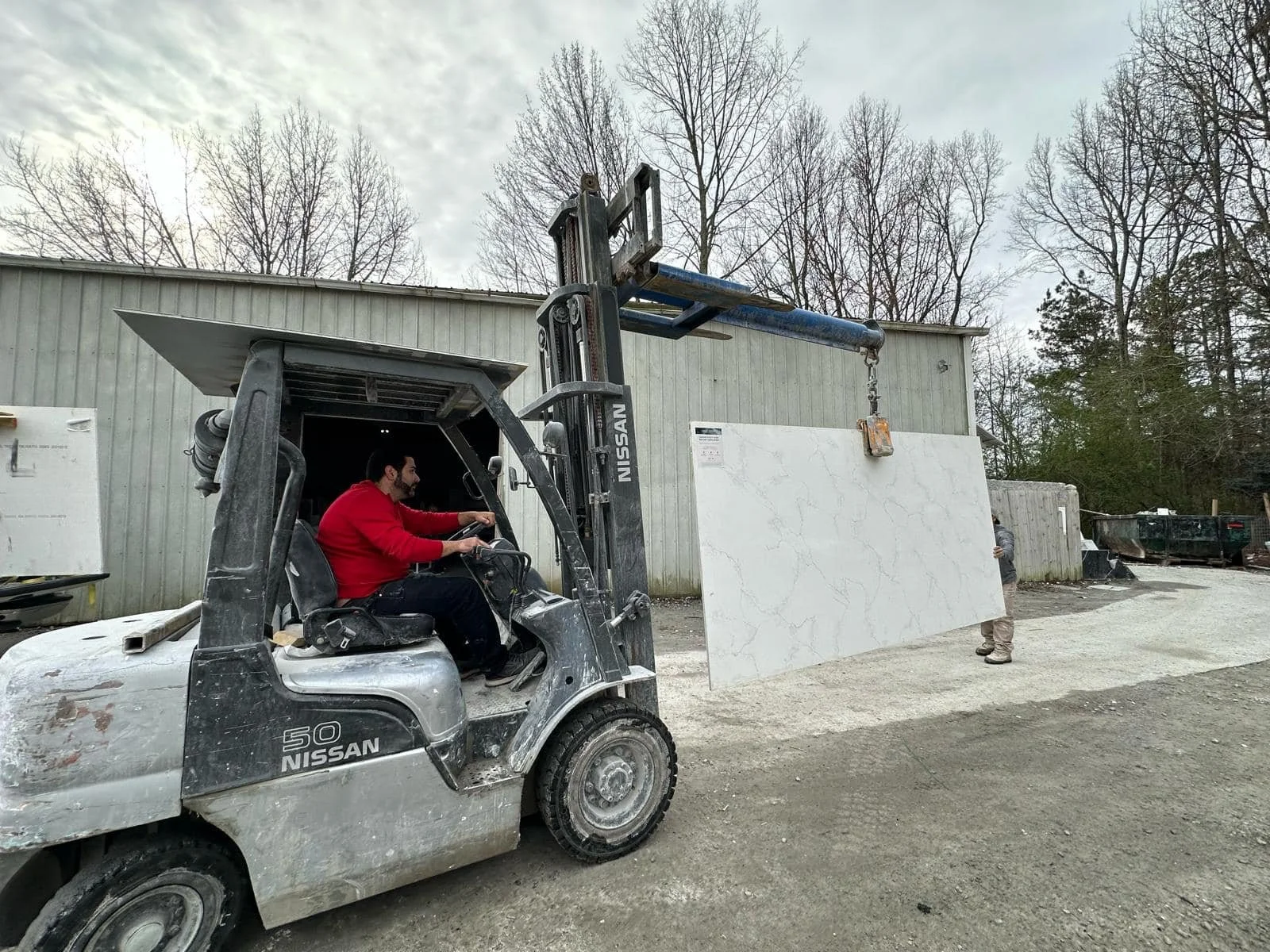 Man operating a forklift pushing a large white marble slab outdoors near a storage building, with another person standing nearby and trees in the background.