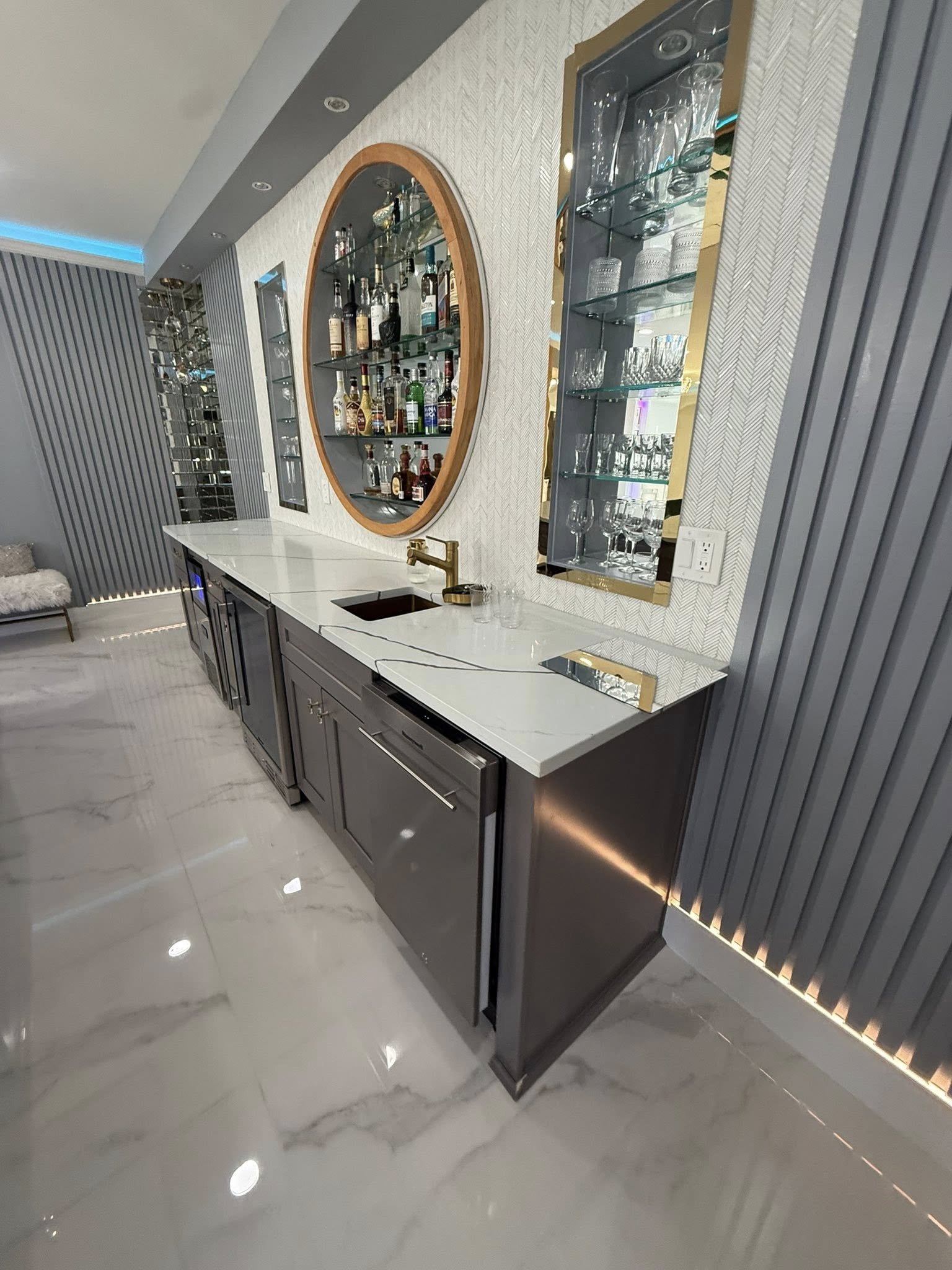 Modern home bar with gray cabinetry, white marble countertop, gold accents, and glass shelves. There are bottles of alcohol, glasses, and a small sink.