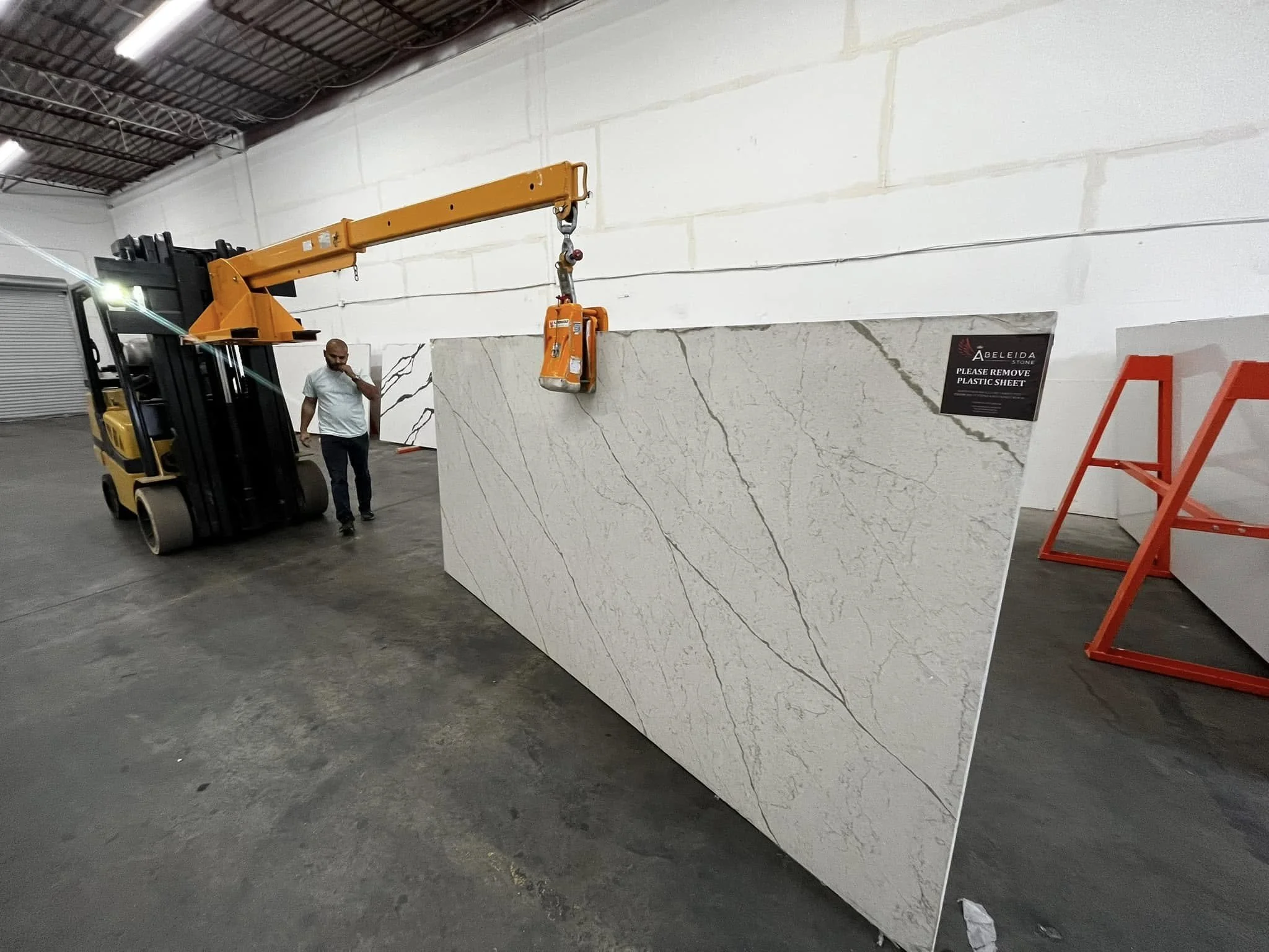 A large piece of white marble with gray veining is being lifted by an orange forklift inside a warehouse. A man in a white shirt and black pants is standing nearby looking at the marble.