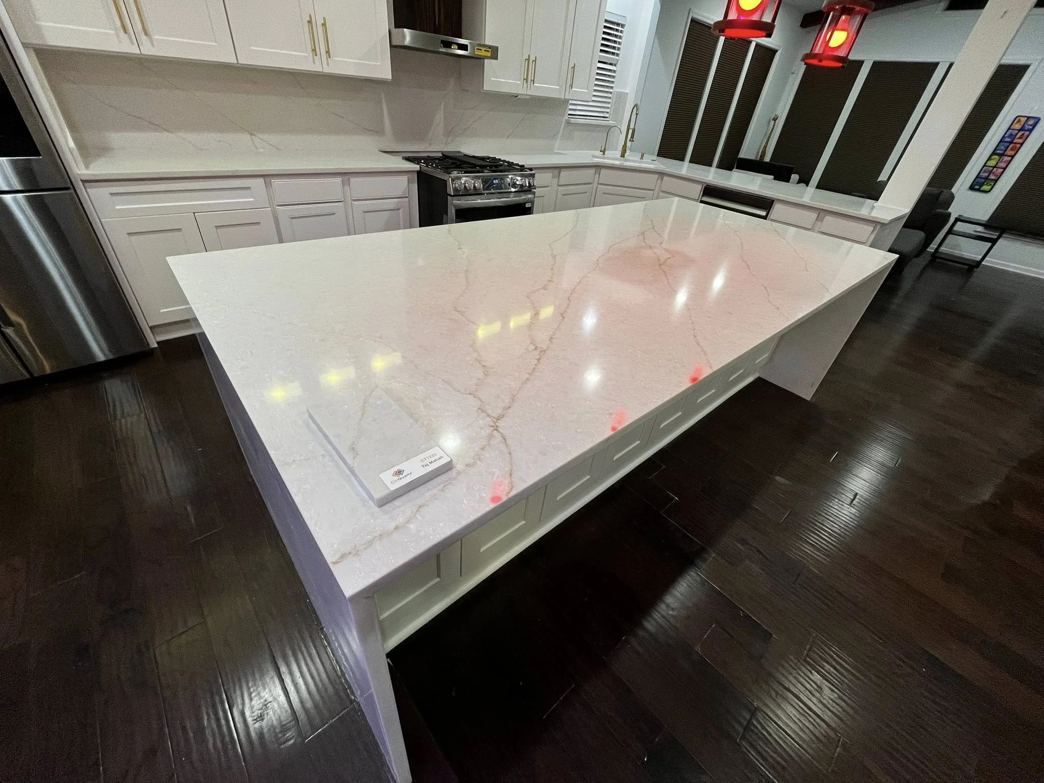 A modern kitchen with white cabinets, a white marble island, stainless steel appliances, dark hardwood floors, and red pendant lights.