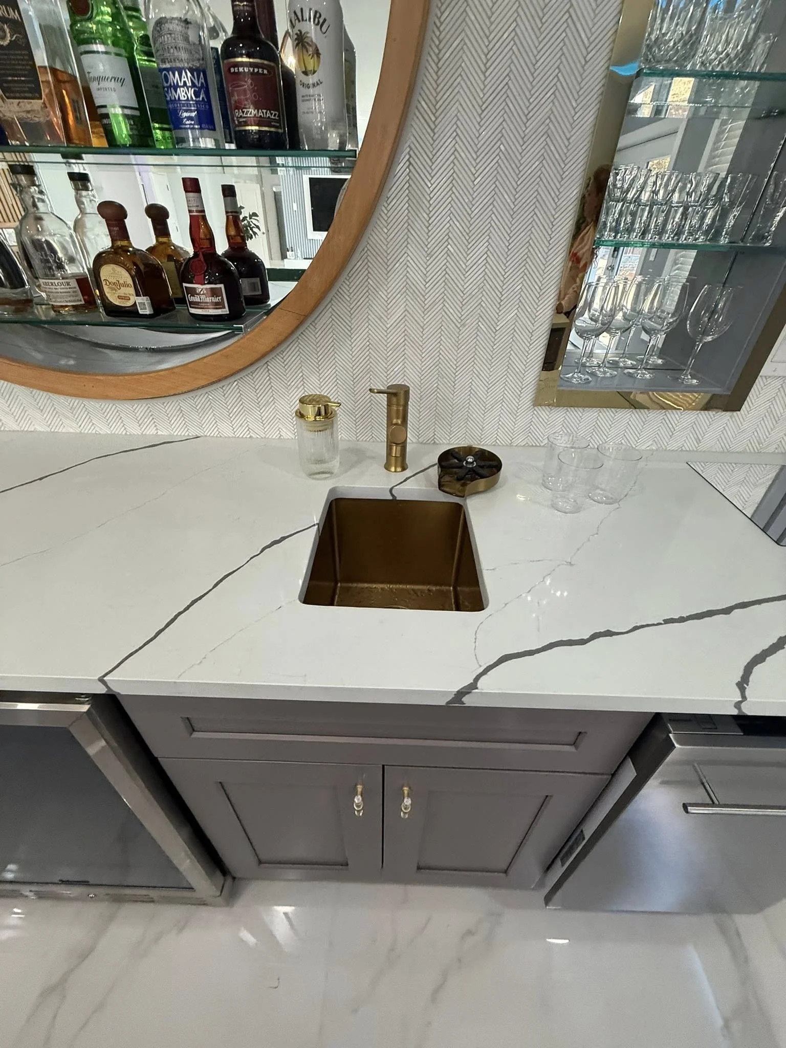Kitchen countertop with a white marble surface featuring dark veining, a small bronze sink, and a gold faucet. Behind the counter is a wall with textured patterned wallpaper, a glass shelf with alcohol bottles including gin and whiskey, and a mirrore