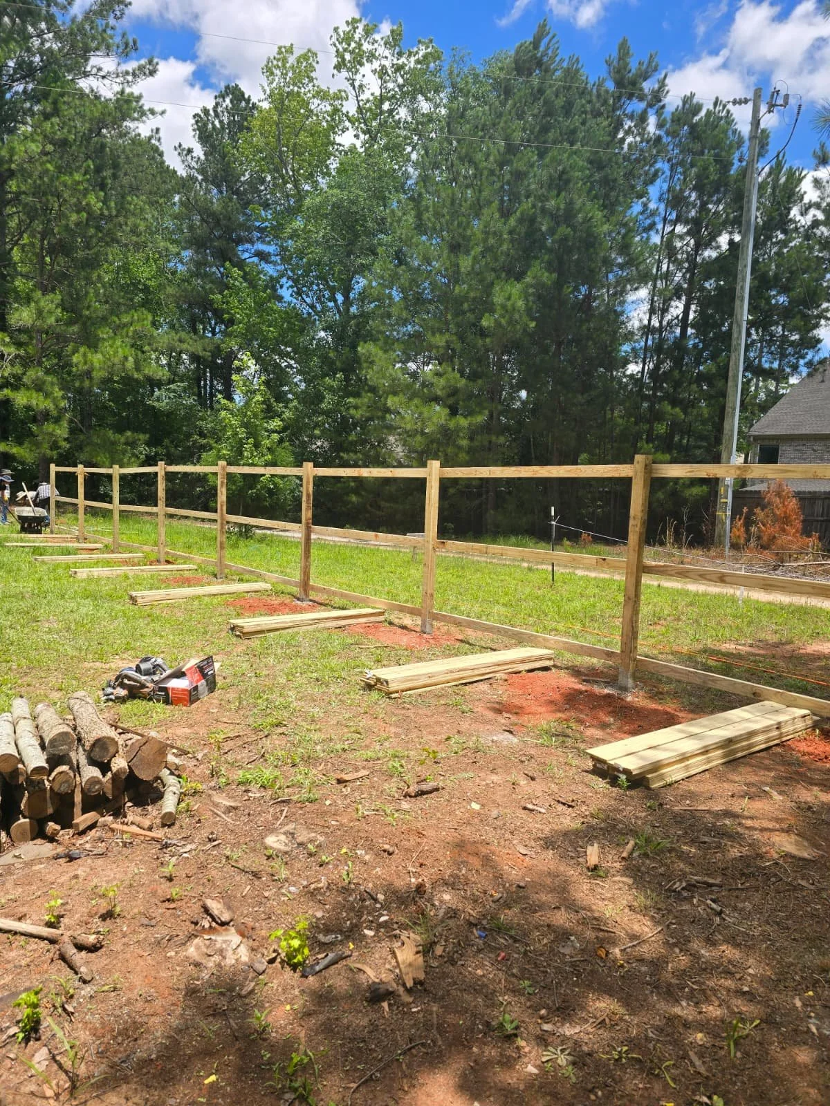 Wooden fence and construction materials in a grassy yard with trees and a cloudy sky in the background.