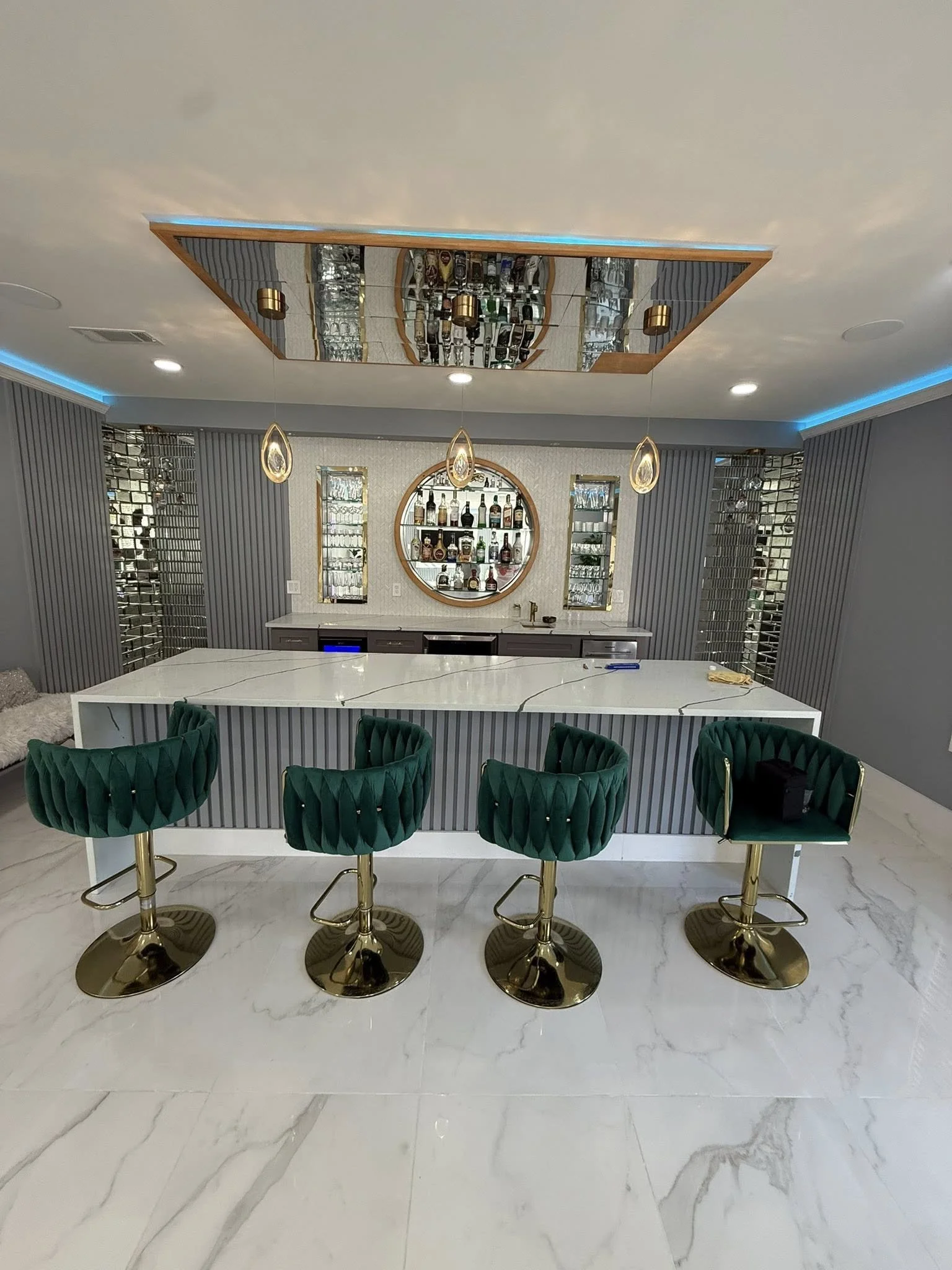 Modern home bar with white marble countertop, four green velvet bar stools with gold bases, mirrored wall with shelves of liquor bottles, and decorative lighting.