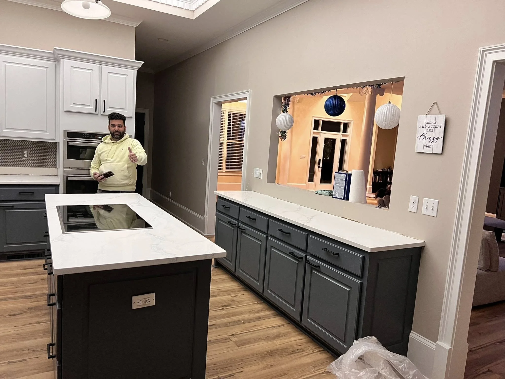 A man giving a thumbs up in a modern kitchen with white and gray cabinets, a white kitchen island with a black stovetop, and a beige wall with decorative hanging lanterns visible through a cutout in the wall.