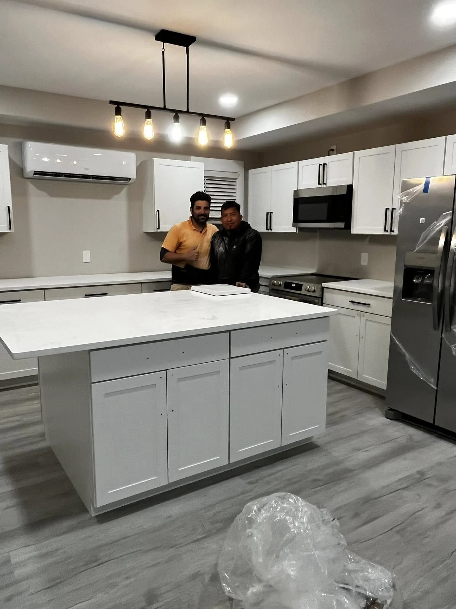Two men standing together in a modern kitchen with white cabinets, gray flooring, and stainless steel appliances, including a refrigerator, microwave, and oven. There is an island in the center and an industrial-style light fixture above.