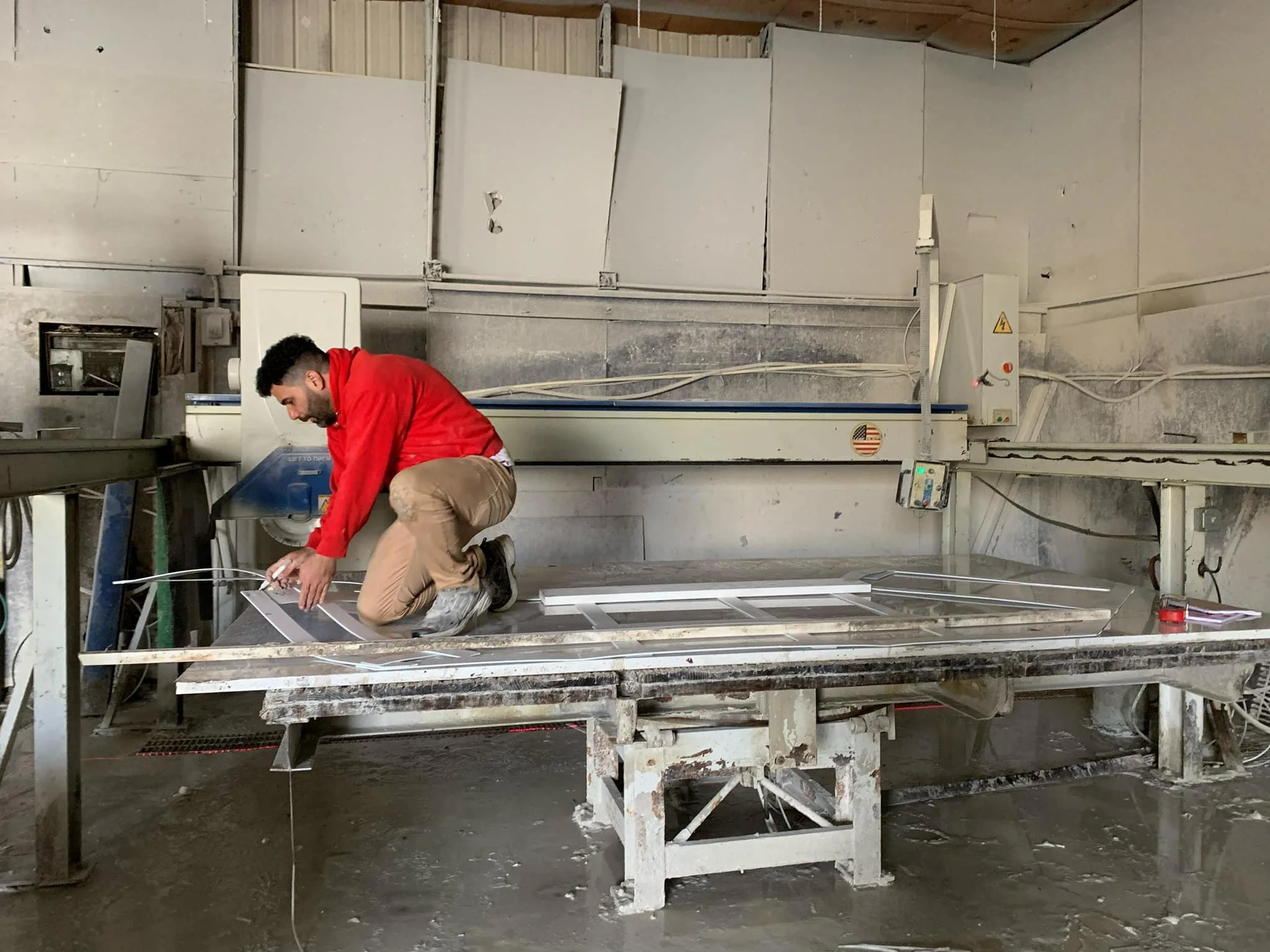 A man in a red shirt and khaki pants kneels on a large industrial glass table in a workshop, working on glass or metal frames. The workshop has a metal and concrete interior with various machinery and tools around.