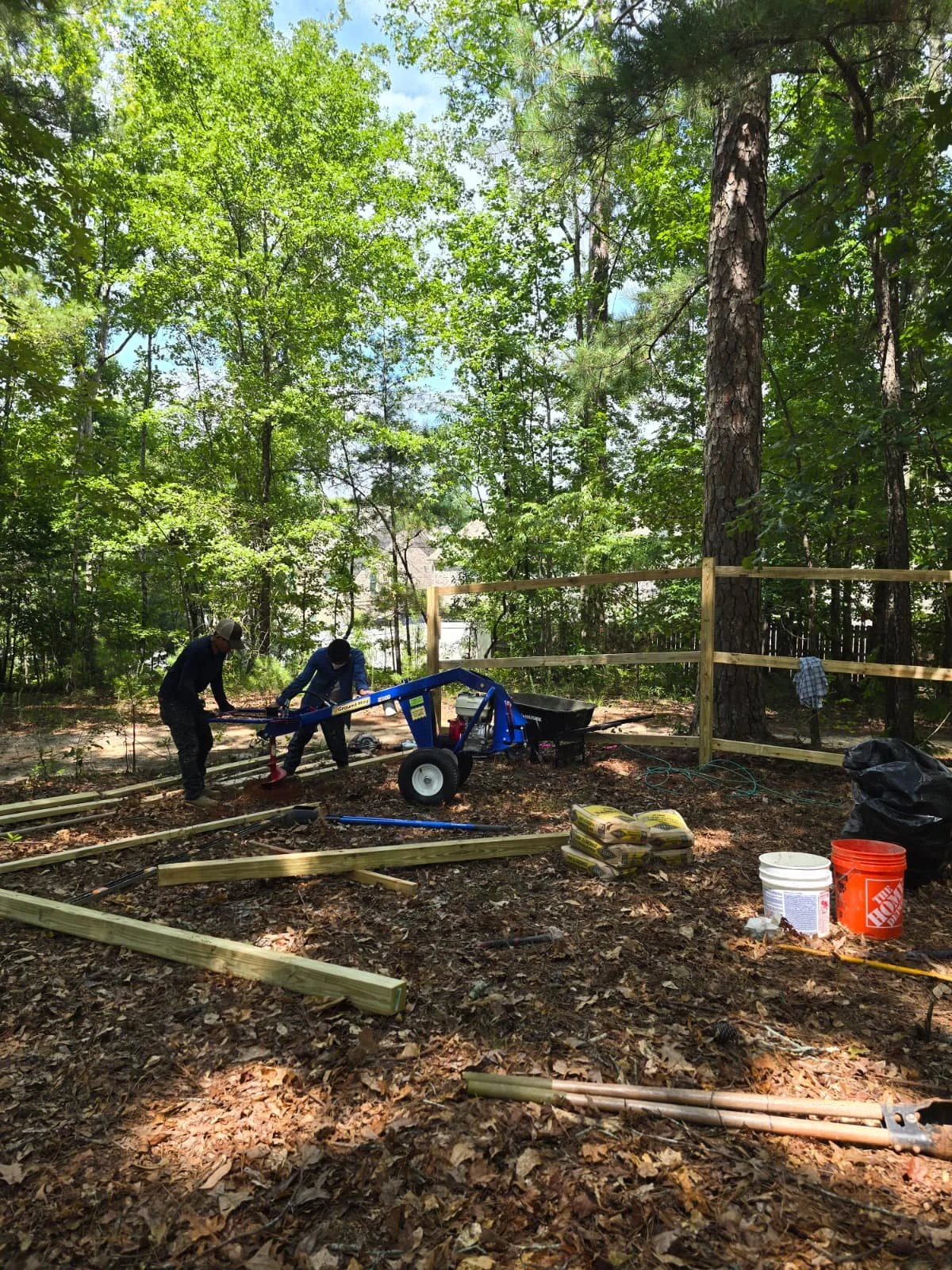 Two men building a wooden fence in a wooded area, with construction supplies, tools, and a blue power auger on the ground.