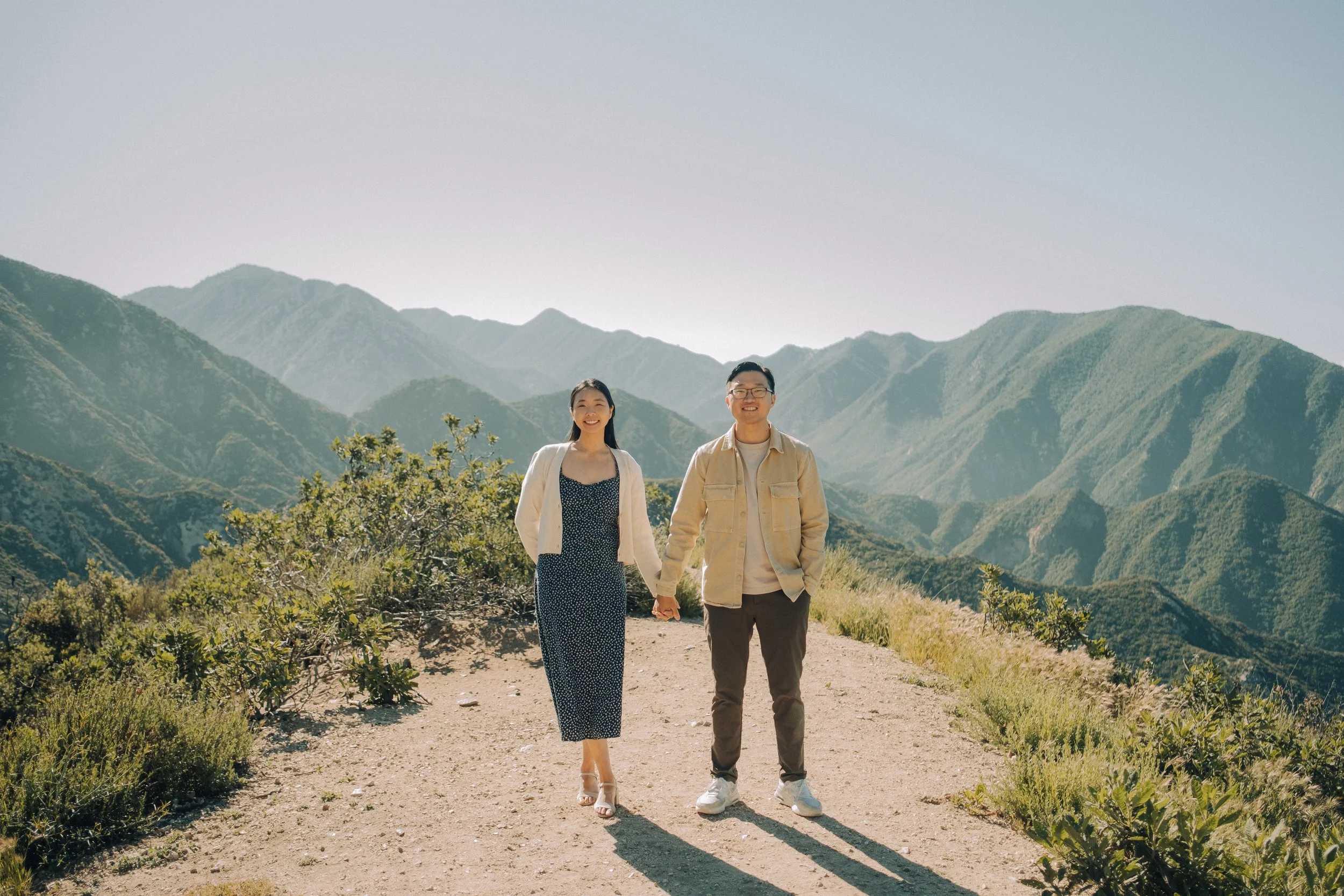 A smiling couple holding hands while walking on a dirt trail in the mountains with lush green peaks and a clear sky in the background.