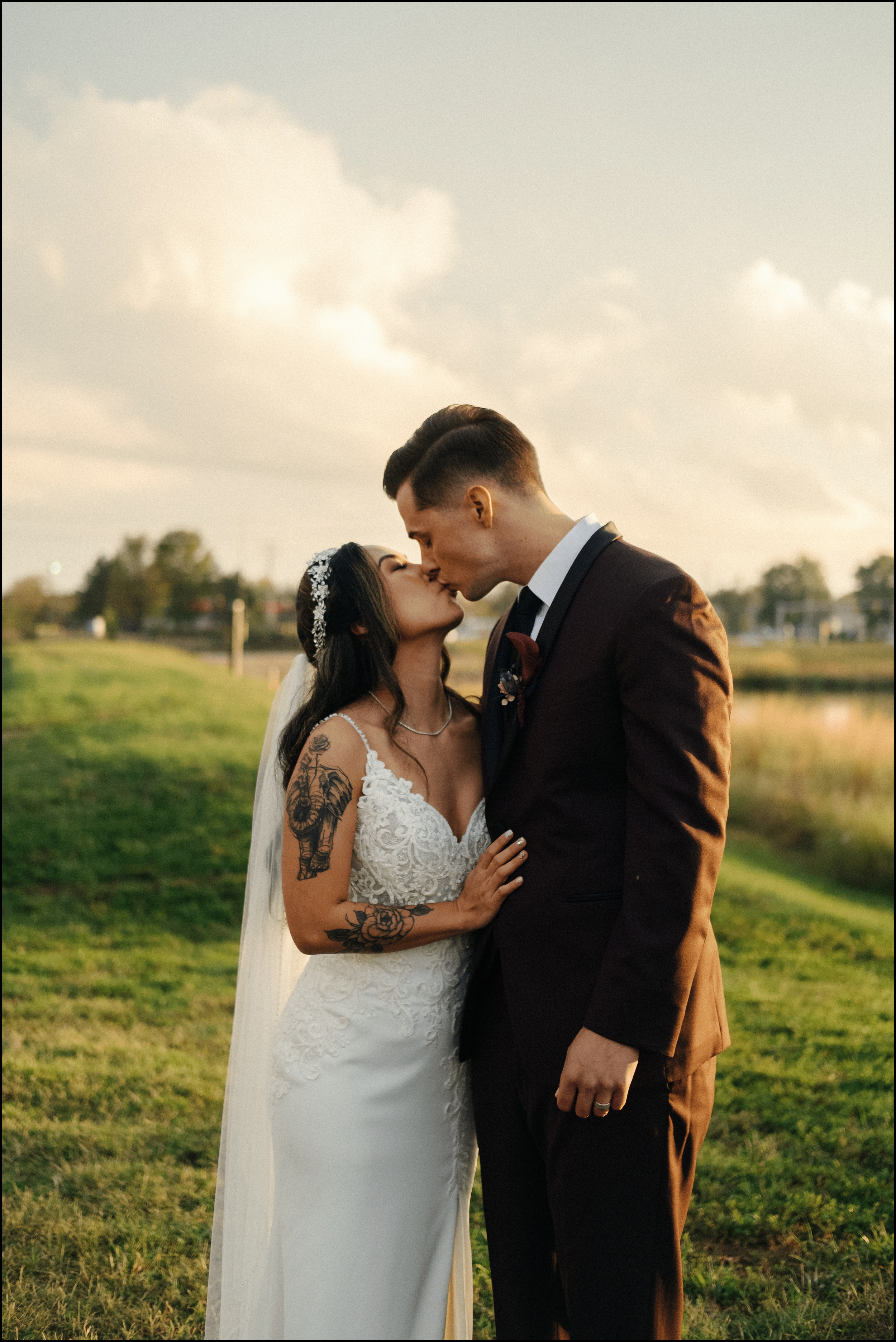 A bride and groom sharing a kiss outdoors during sunset, with trees, grass, and a small body of water in the background.