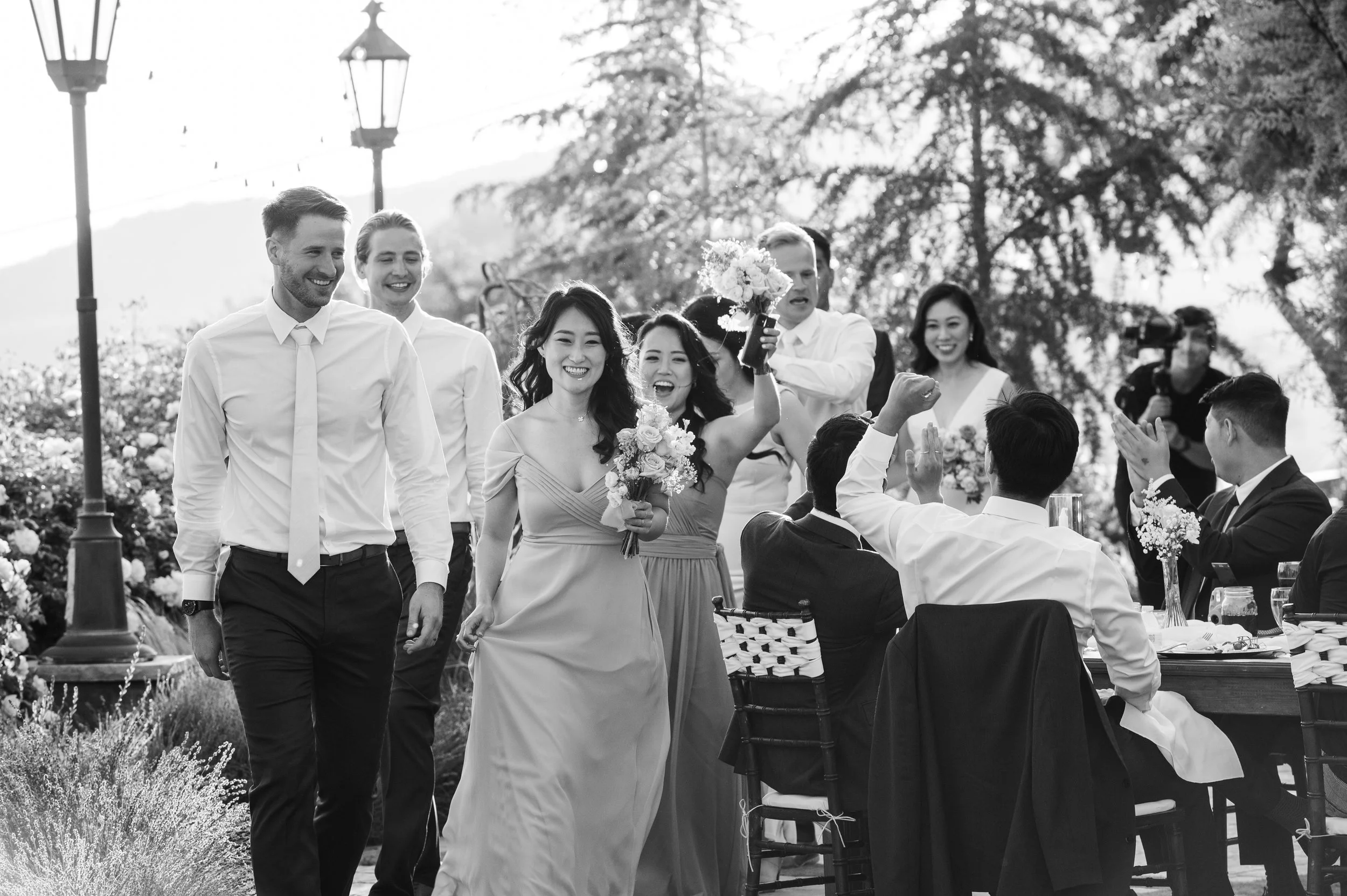 Group of people at a wedding celebration outdoors, with some sitting at a banquet table and others standing, smiling, and holding flowers, with trees and decorative lampposts in the background.