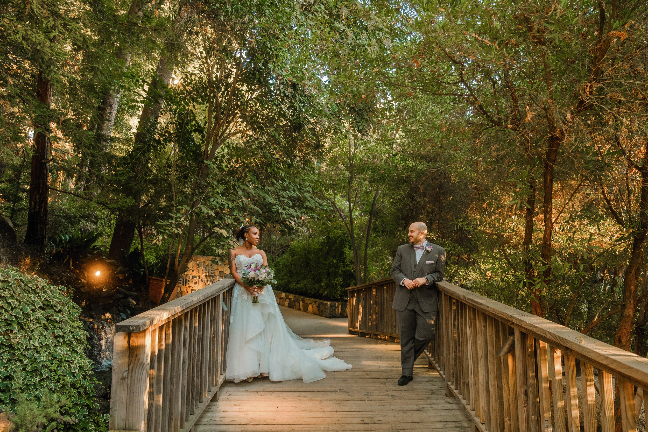 A bride and groom stand on a wooden bridge surrounded by lush greenery, with the bride holding a bouquet and the groom leaning casually on the railing, both smiling at each other during their wedding photos.