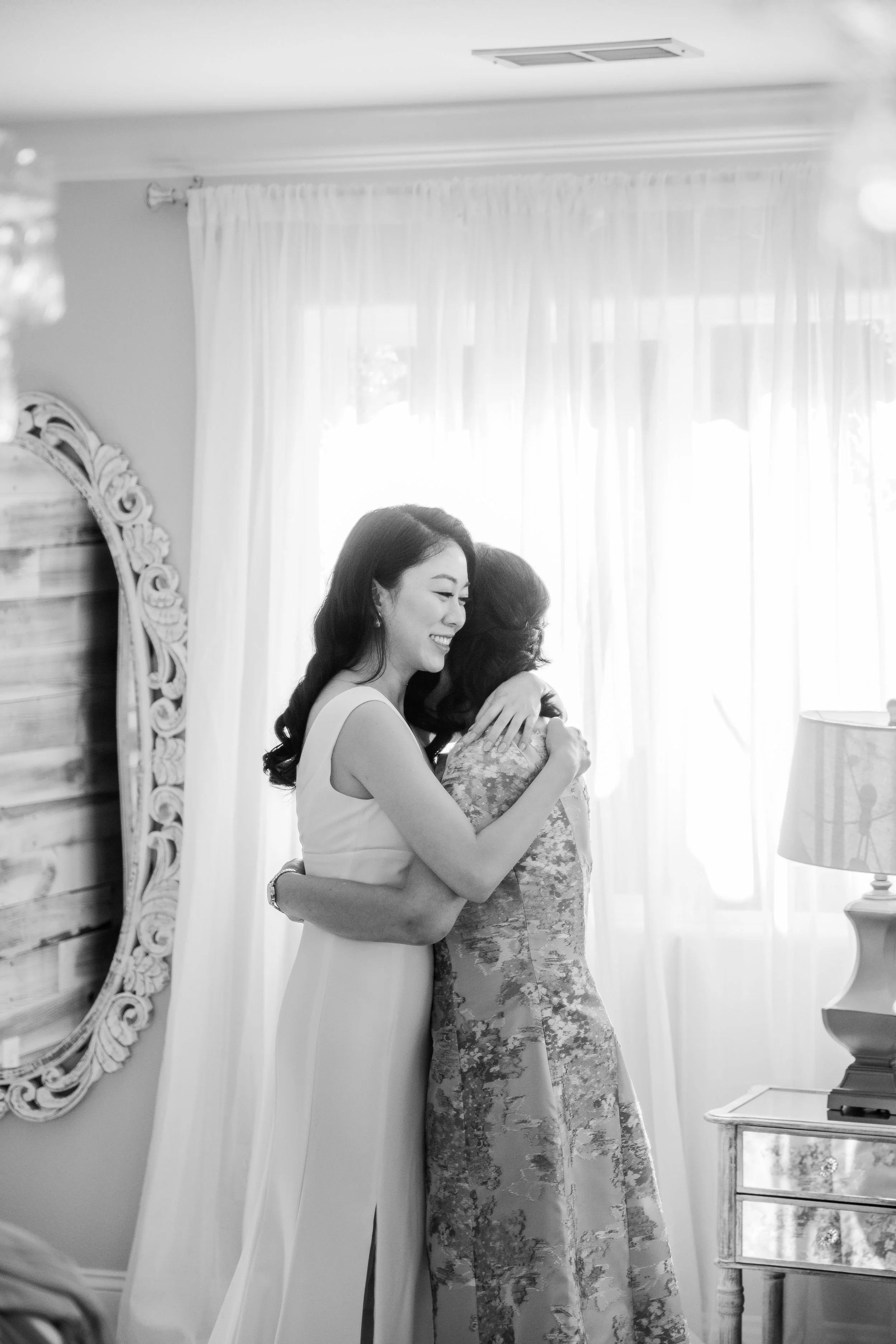 Two women embracing, one in a white dress hugging another in a patterned dress, inside a bright room with curtains and a mirror.