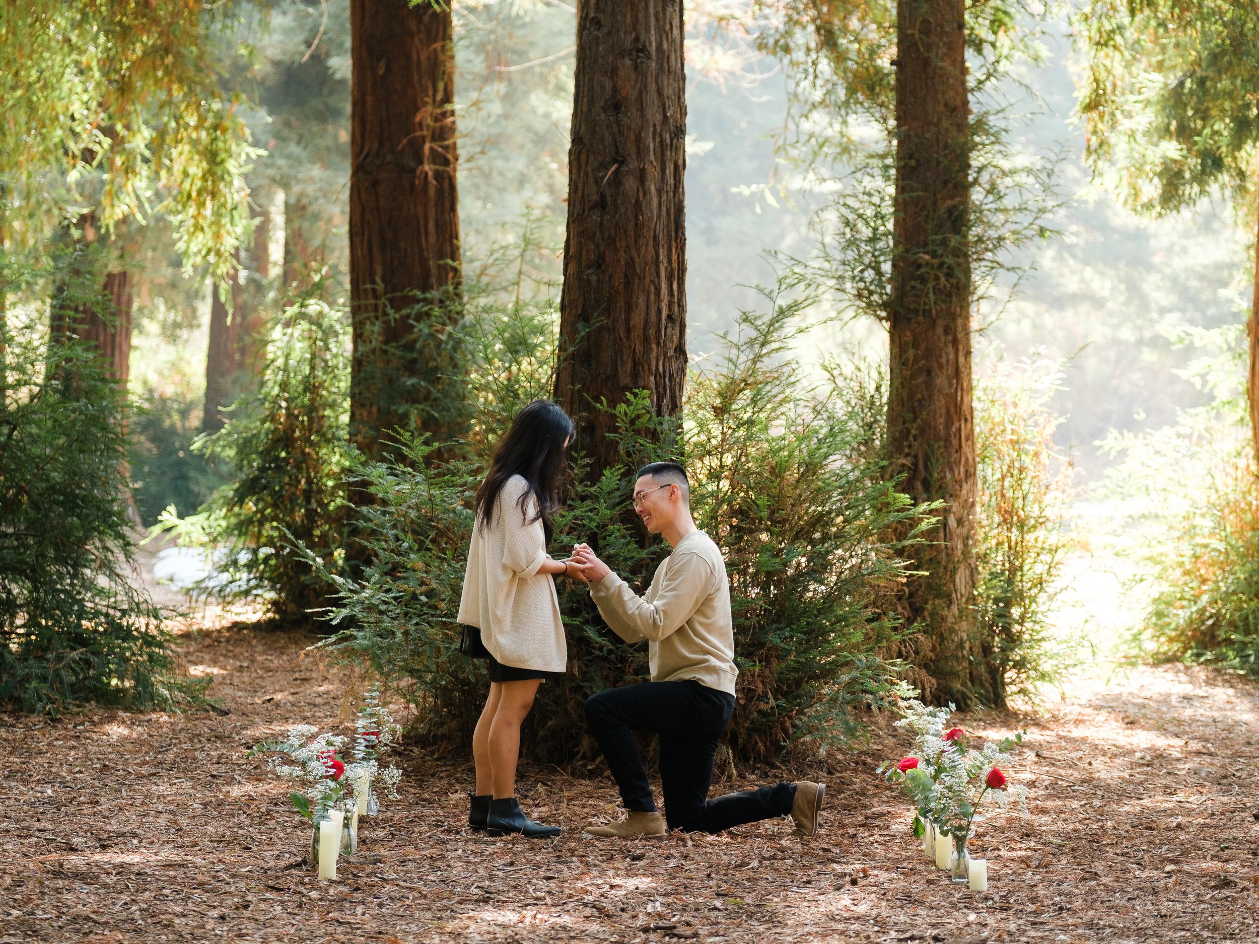 A man proposes marriage to a woman in a forest, kneeling on one knee while holding her hands. The woman is standing and looking at him, and there are flowers and candles arranged on the ground around them.