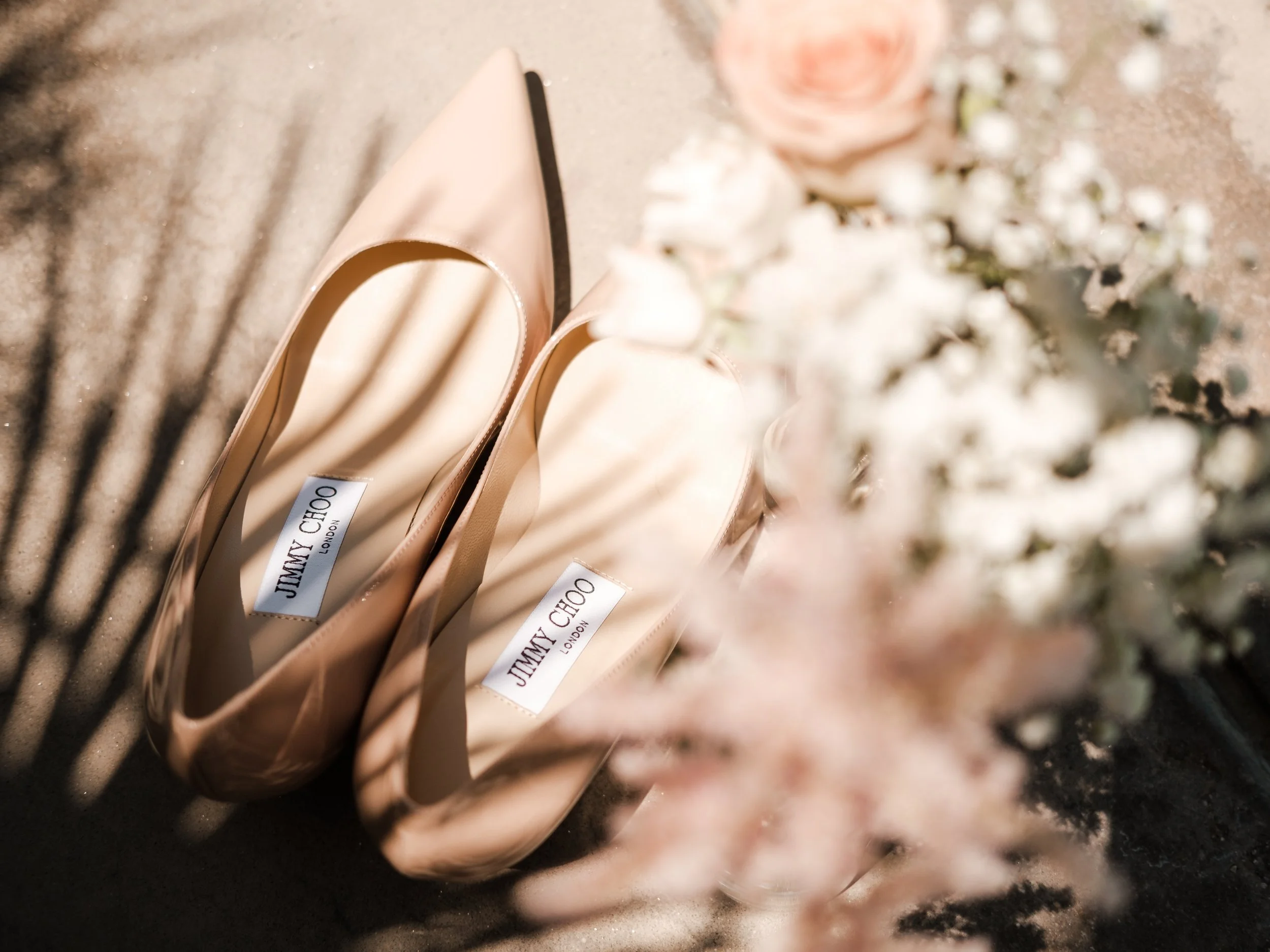Beige Jimmy Choo high-heeled shoes placed on a textured surface with shadows and some white flowers in the foreground.