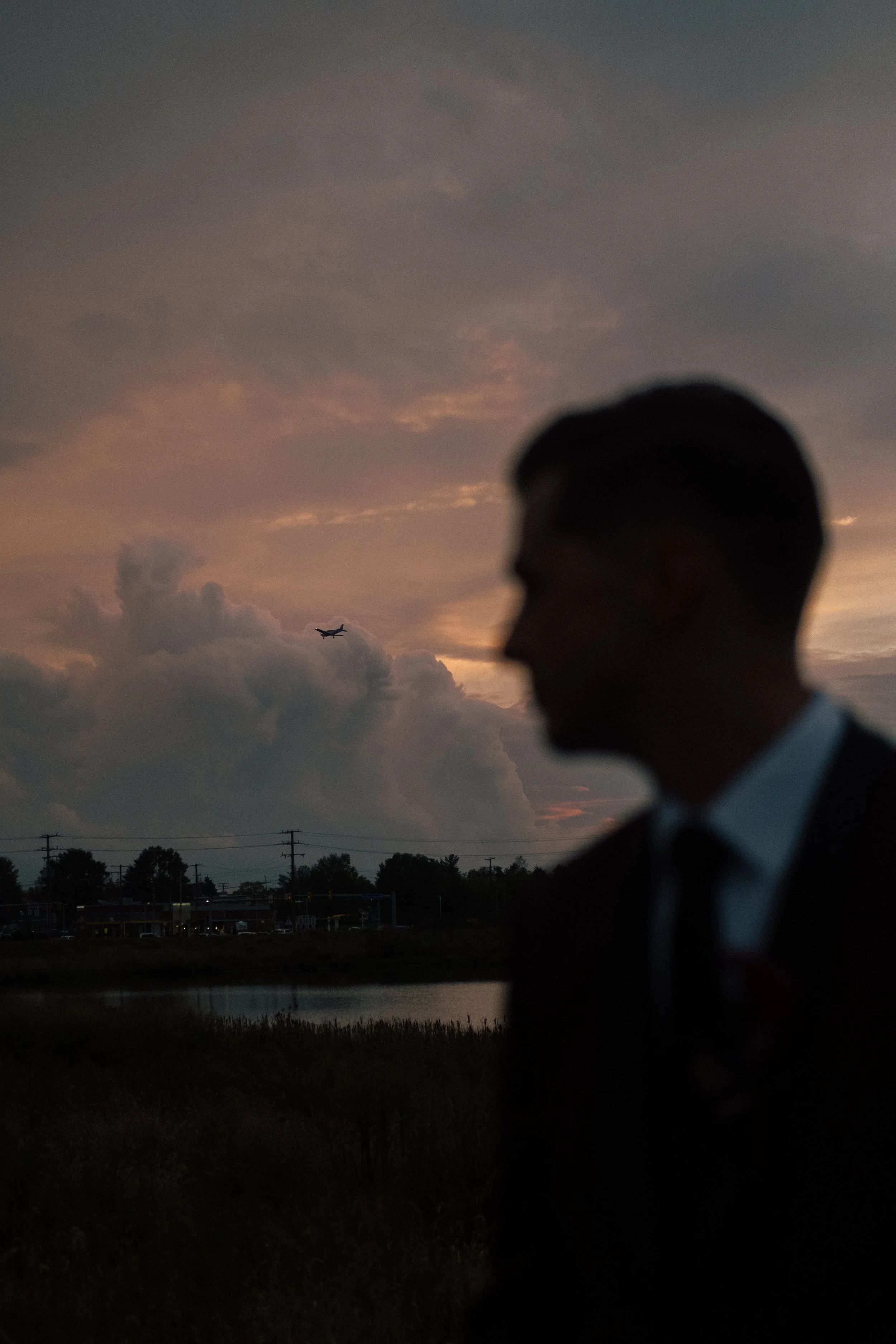 Silhouette of a man in a suit against a sunset sky with clouds and an airplane in the background.
