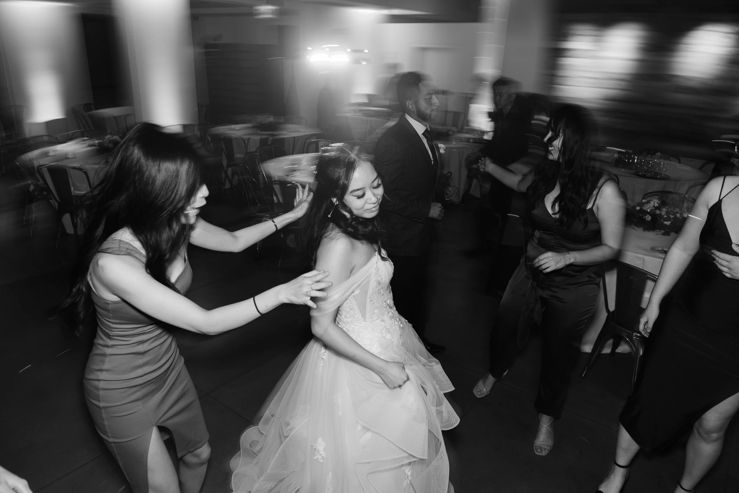A group of people dancing at a wedding reception, including a bride in a wedding gown and several women in dresses, with chairs and tables in the background.