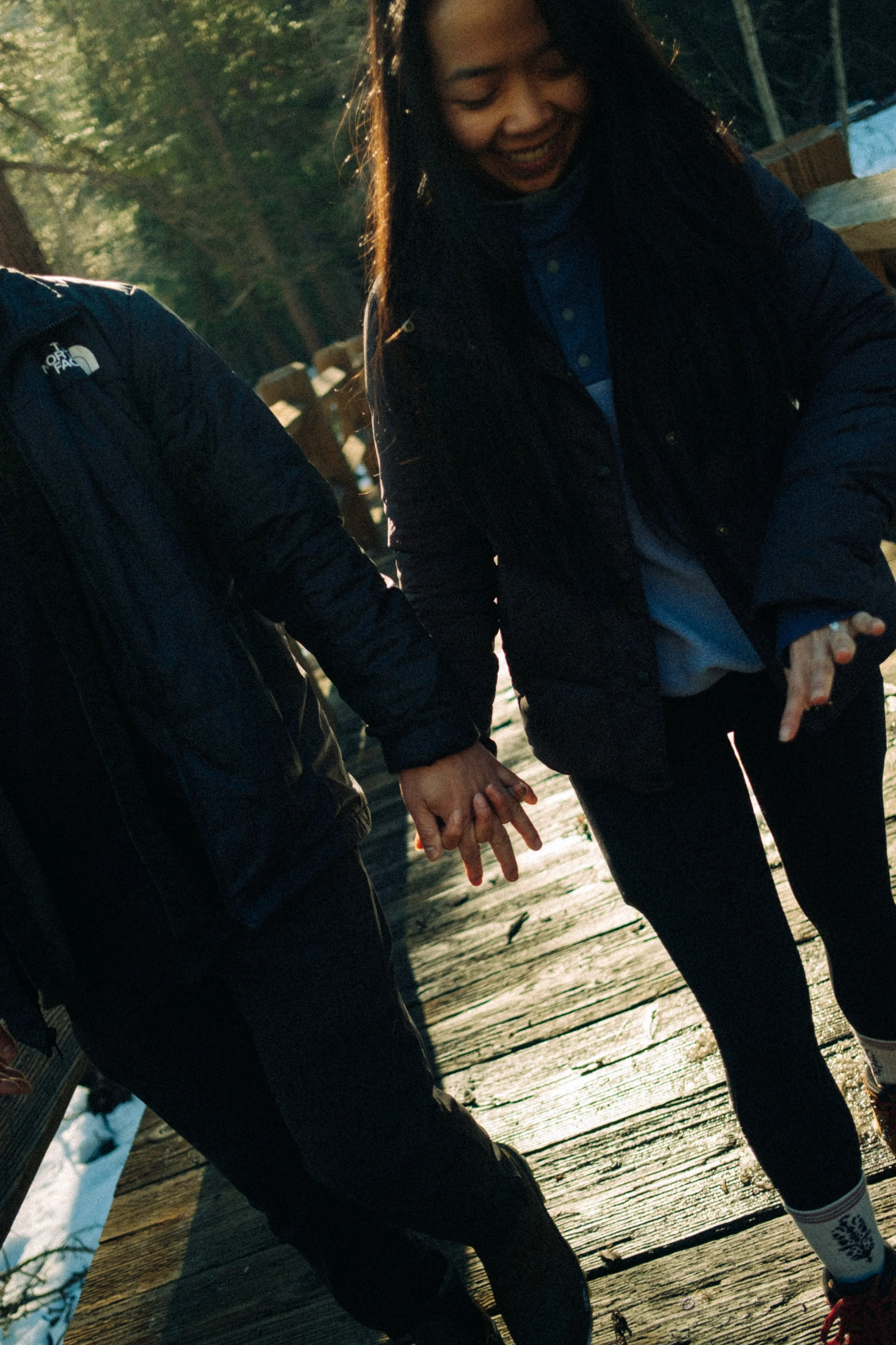 A woman and a person holding hands while walking on a wooden bridge outdoors during daytime.