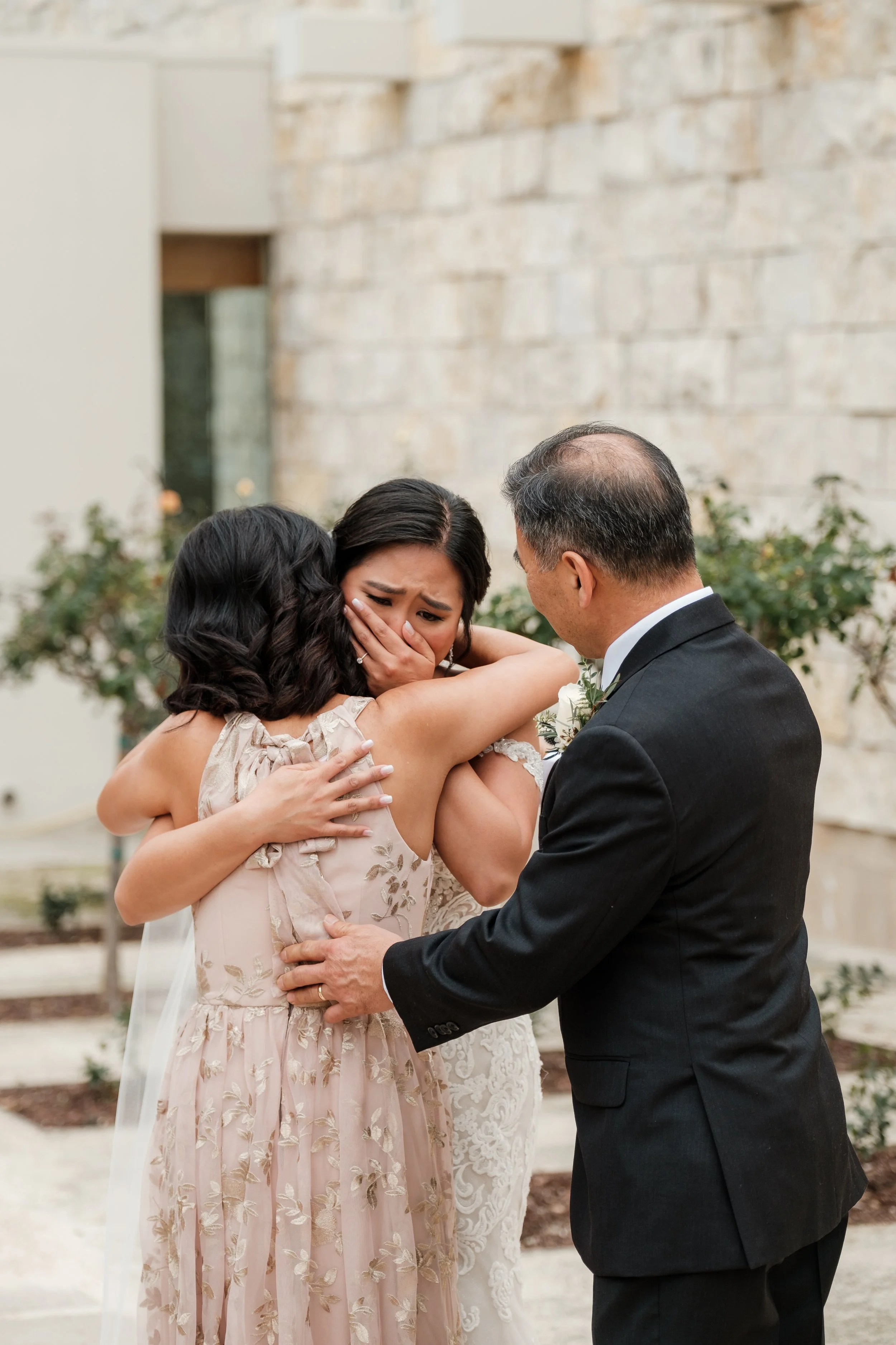 A woman dressed in a wedding gown is crying as she embraces another woman and a man, possibly her parents, during a wedding ceremony outdoors.