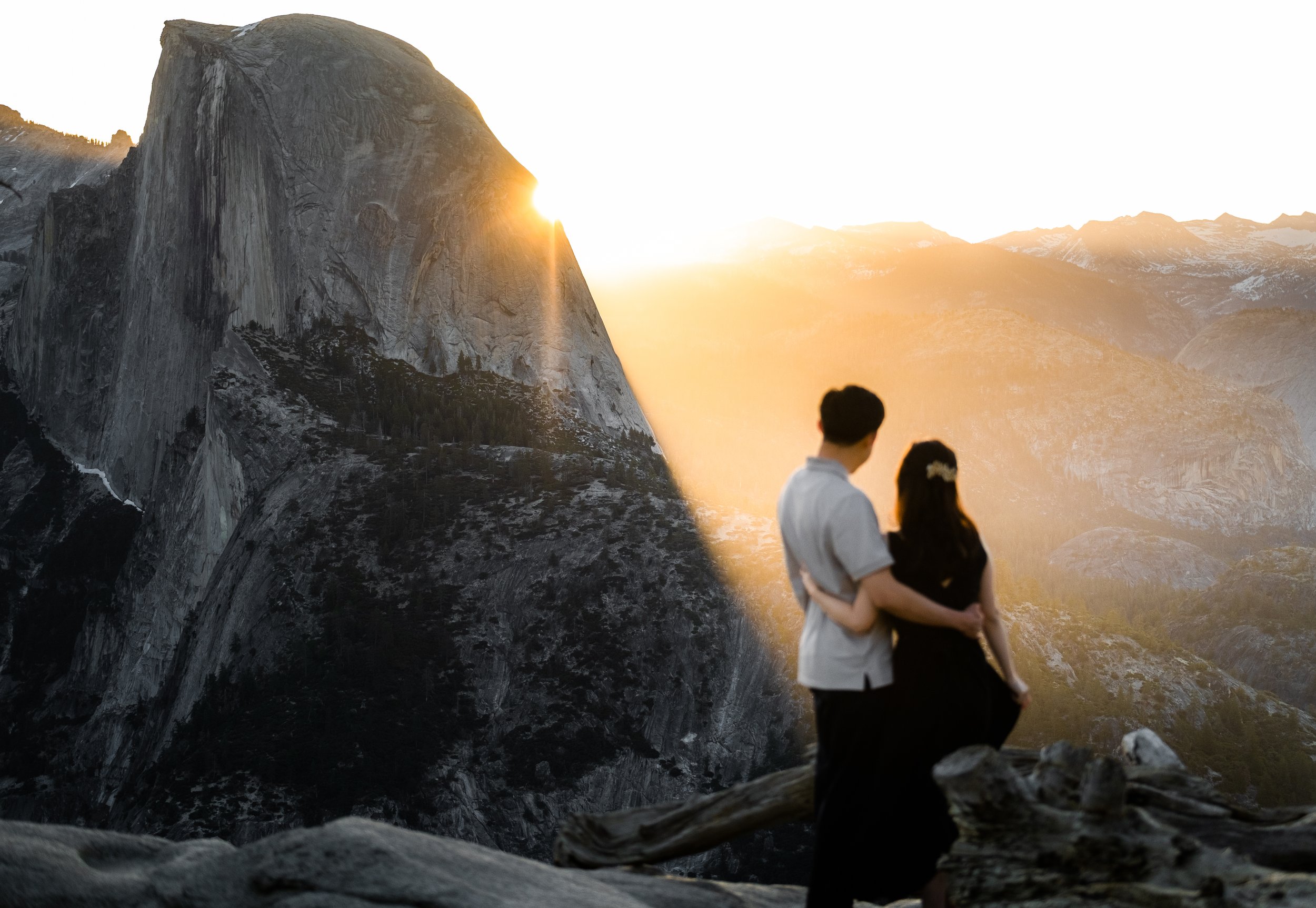 A couple standing on rocks, embracing and looking at a mountain landscape during sunset, with sunlight peeking through the mountains.