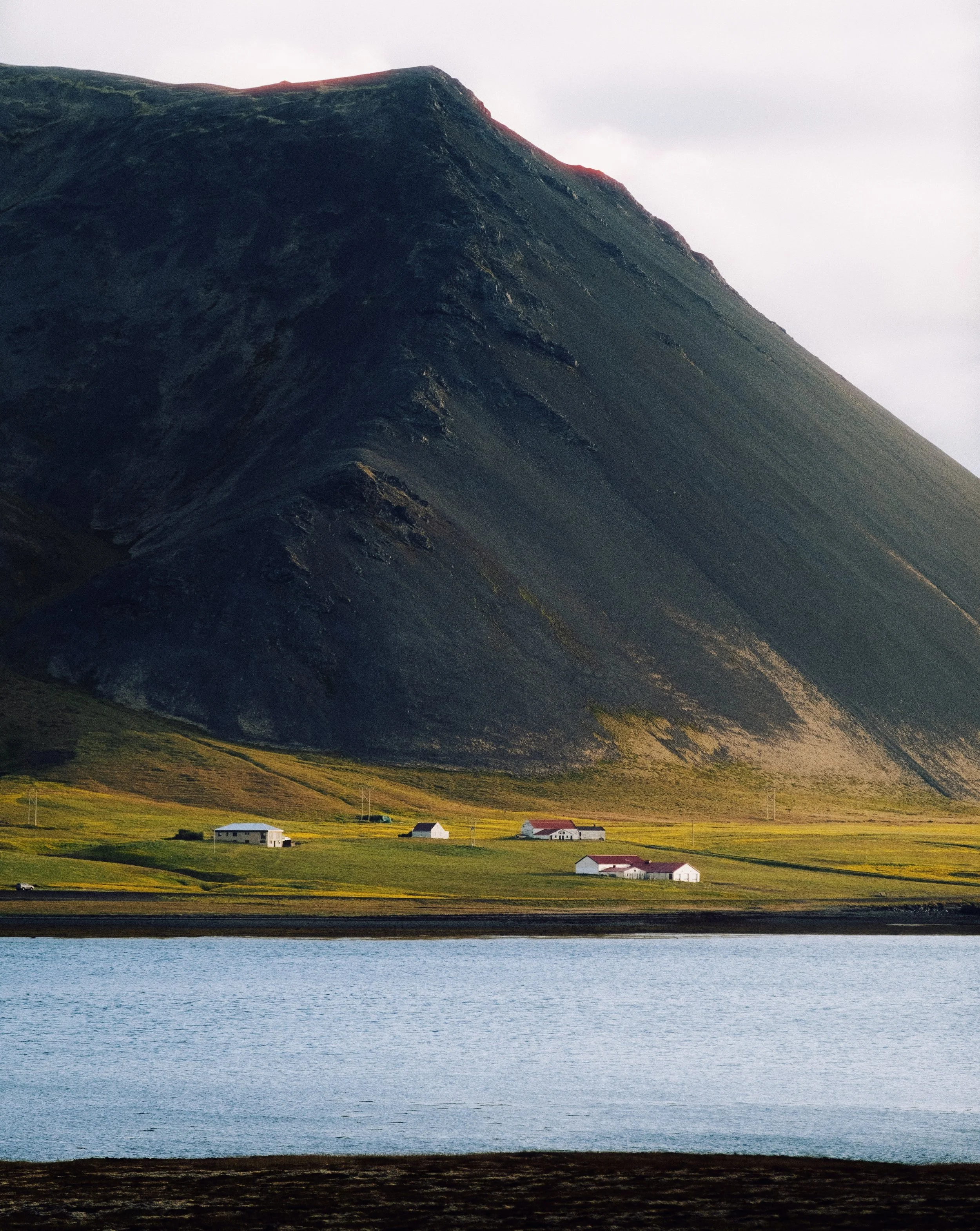 A large mountain with dark, steep slopes and a small cluster of white buildings with red roofs at its base, situated near a body of water.