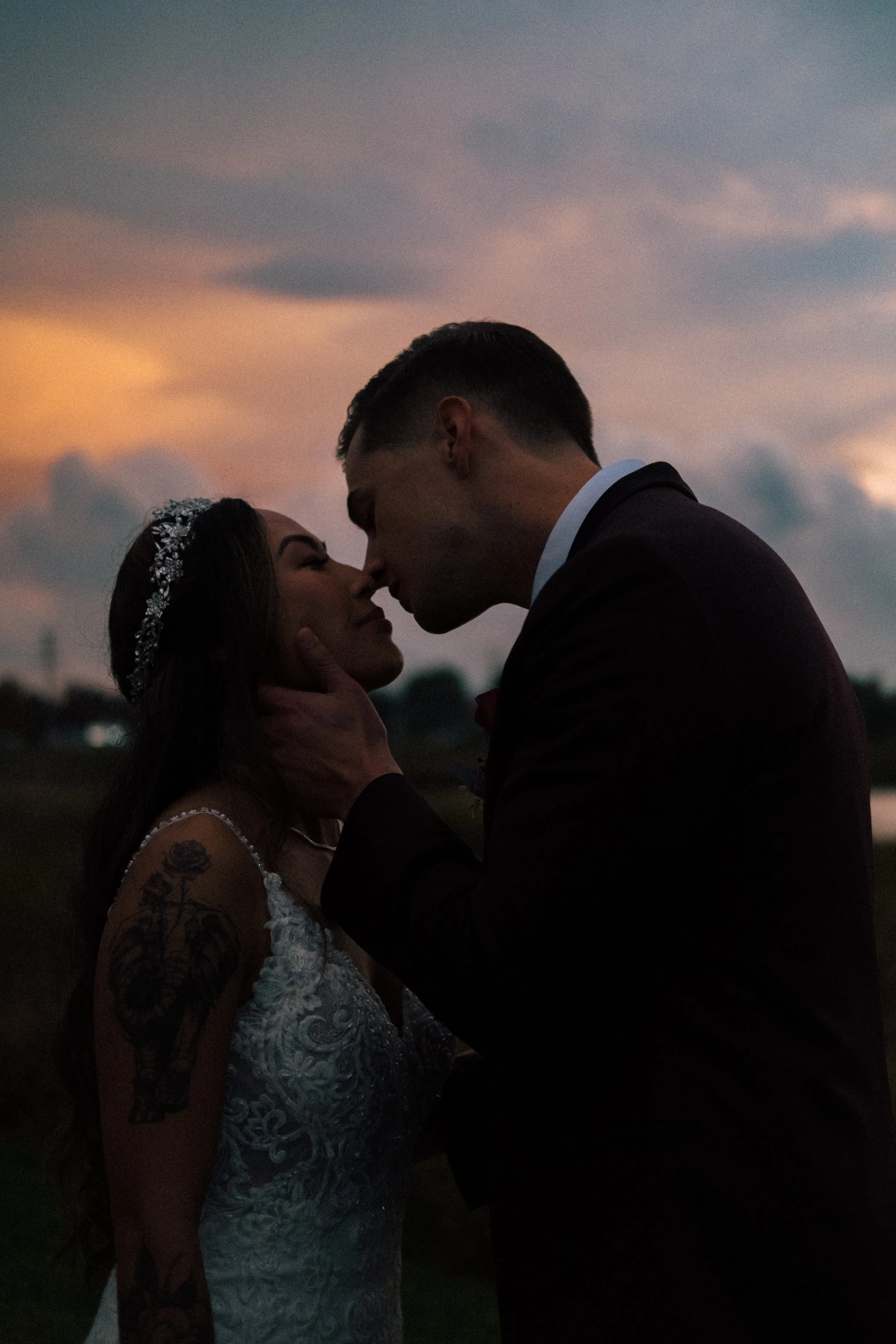 A romantic moment between a bride and groom at sunset, with the groom gently holding the bride's face as they touch noses. The bride is wearing a white wedding dress and a decorative headband, and has tattoos on her arm. The sky is cloudy with warm hues from the setting sun.