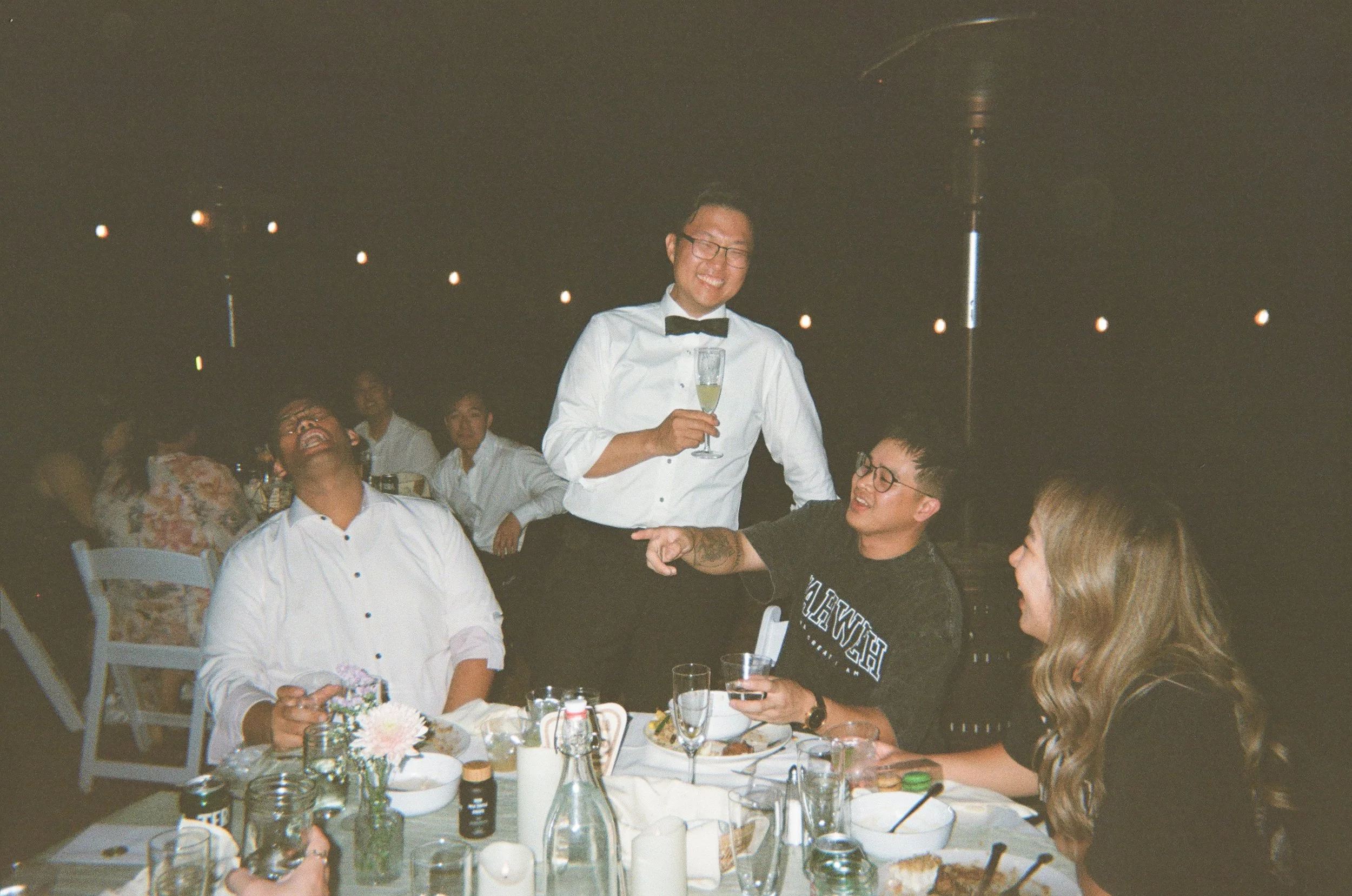 People dining at a restaurant, with one person in a tuxedo holding a glass and others laughing and talking around a table.