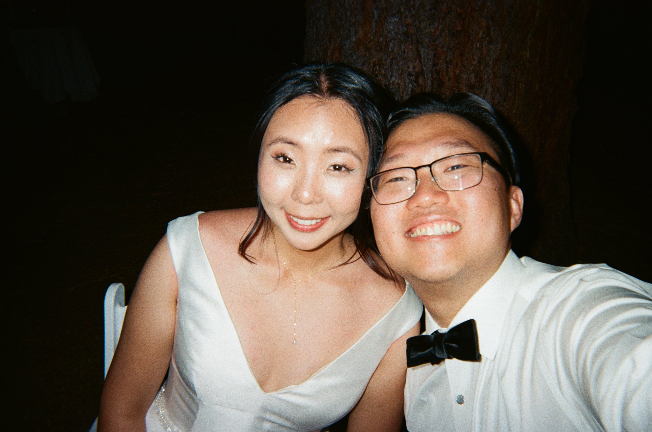 A smiling couple taking a selfie at a formal event, with a woman in a white dress and a man in a white shirt with a black bow tie.