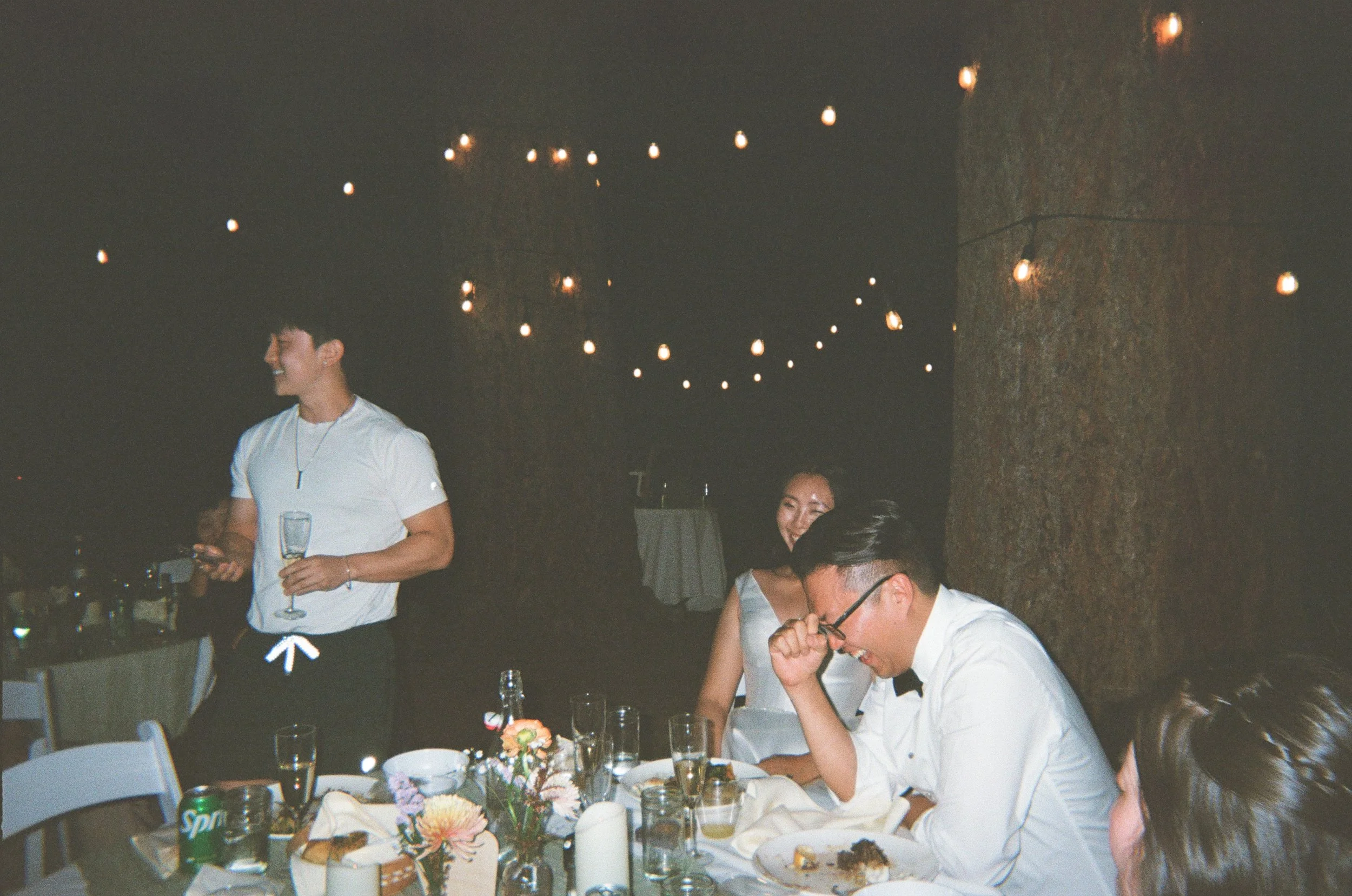 People celebrating at a party with string lights overhead, seated at a table with food and drinks, including a Sprite can, in a dimly lit environment.