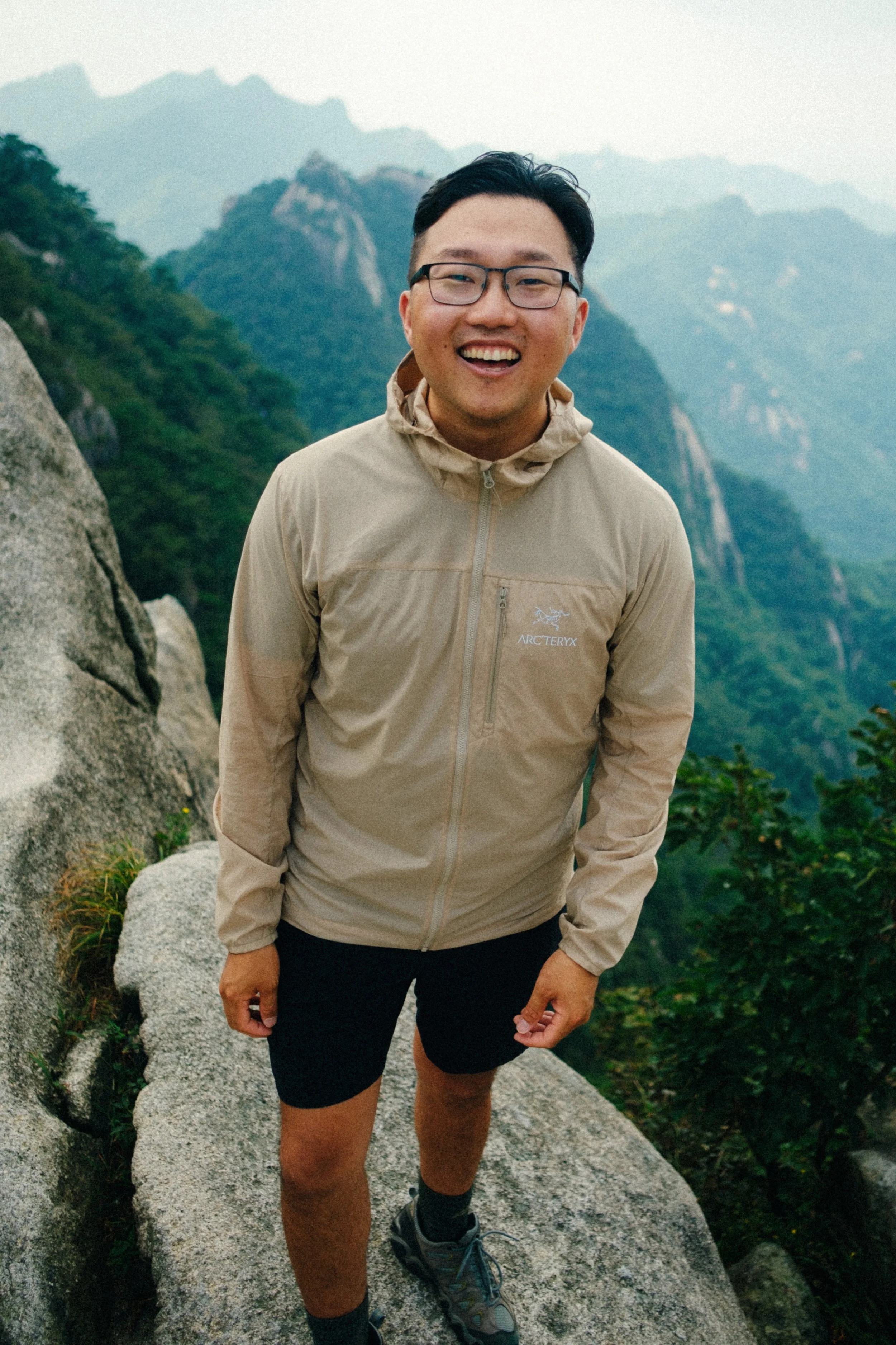 A man smiling and posing outdoors on a mountain trail, wearing a beige Arc'teryx jacket, black shorts, and hiking shoes, with mountain peaks and greenery in the background.