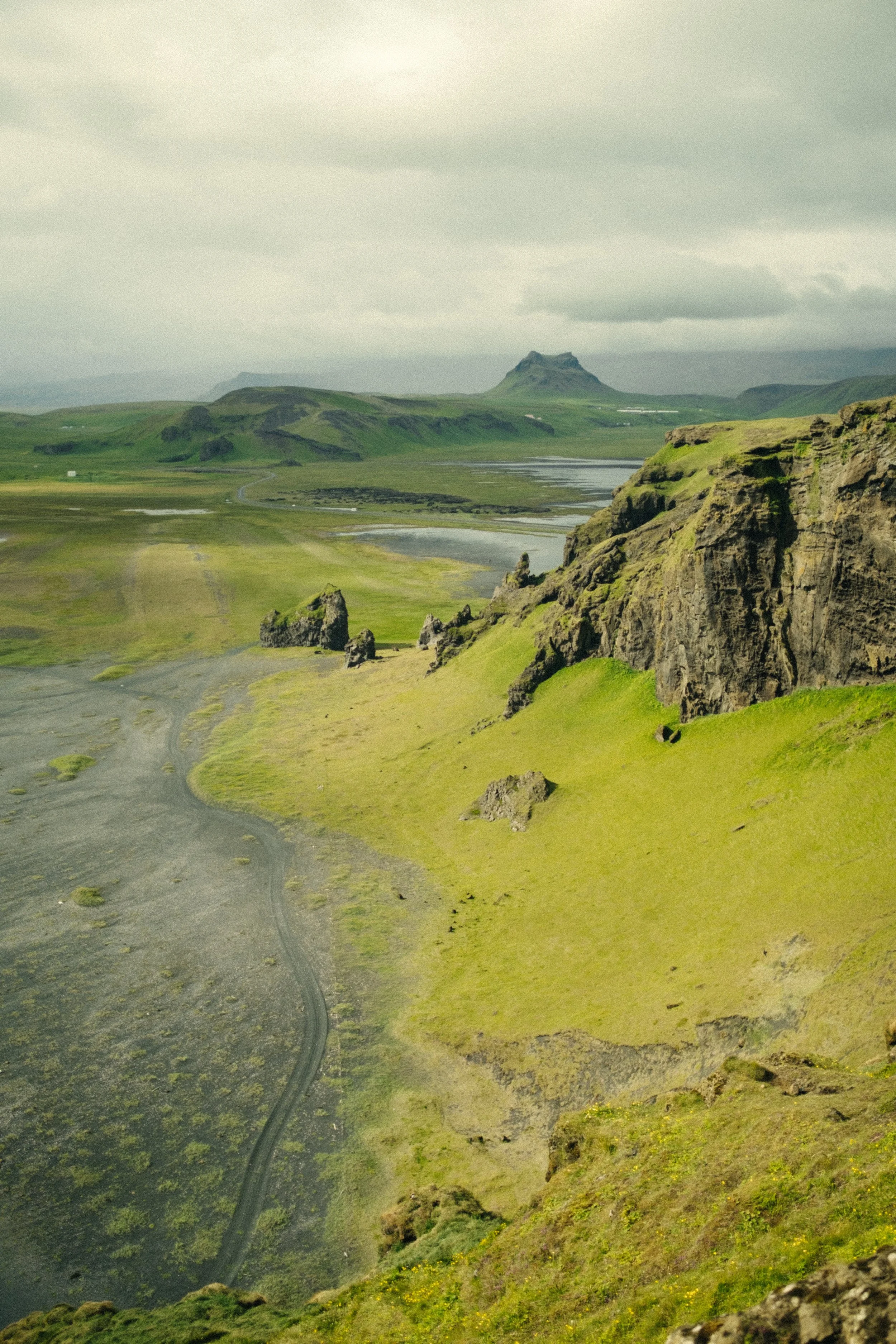 Scenic Icelandic landscape with green hills, rocky cliffs, a winding dirt road, and distant mountains under cloudy sky.