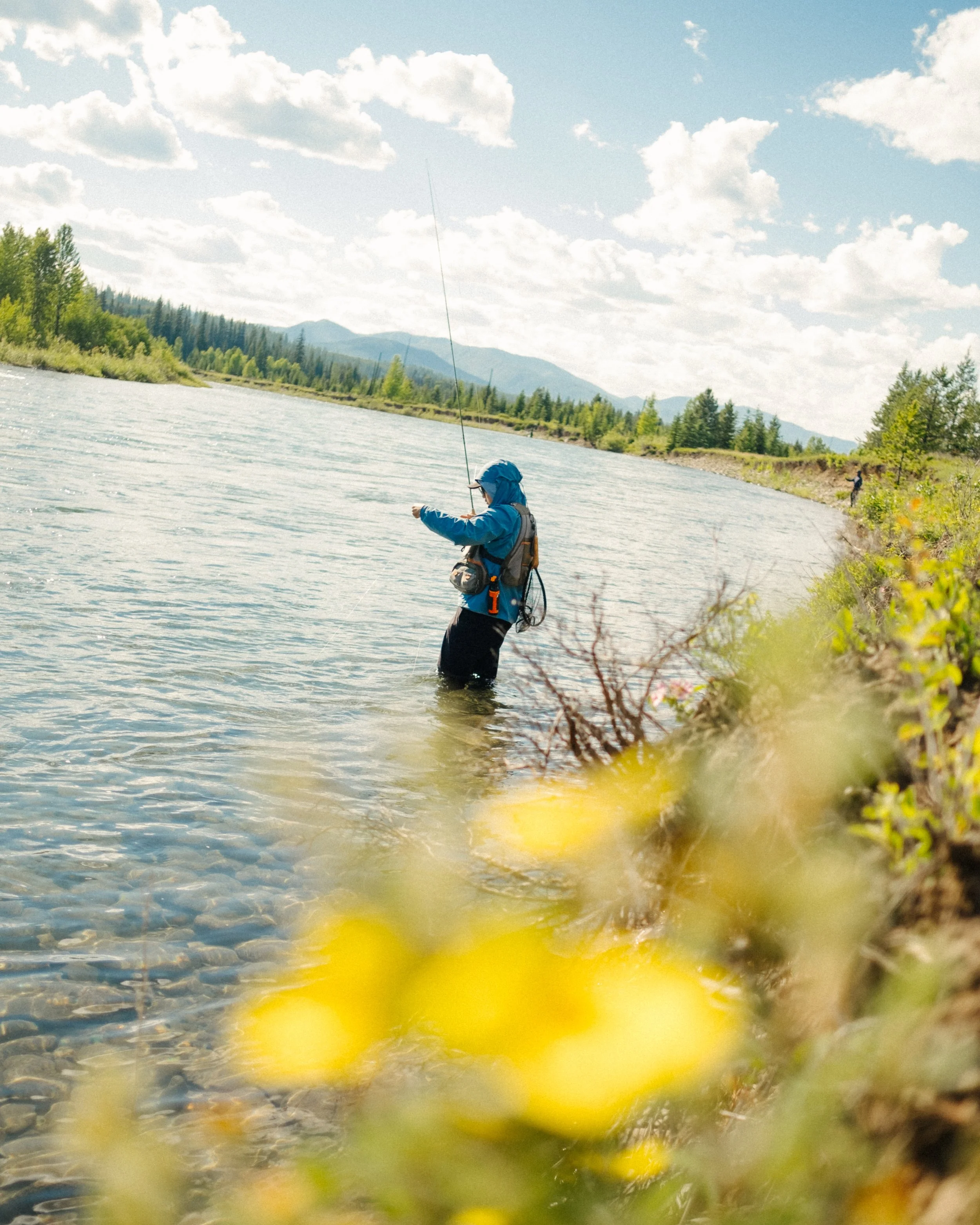 Person fishing in a river with mountains and blue sky in the background.