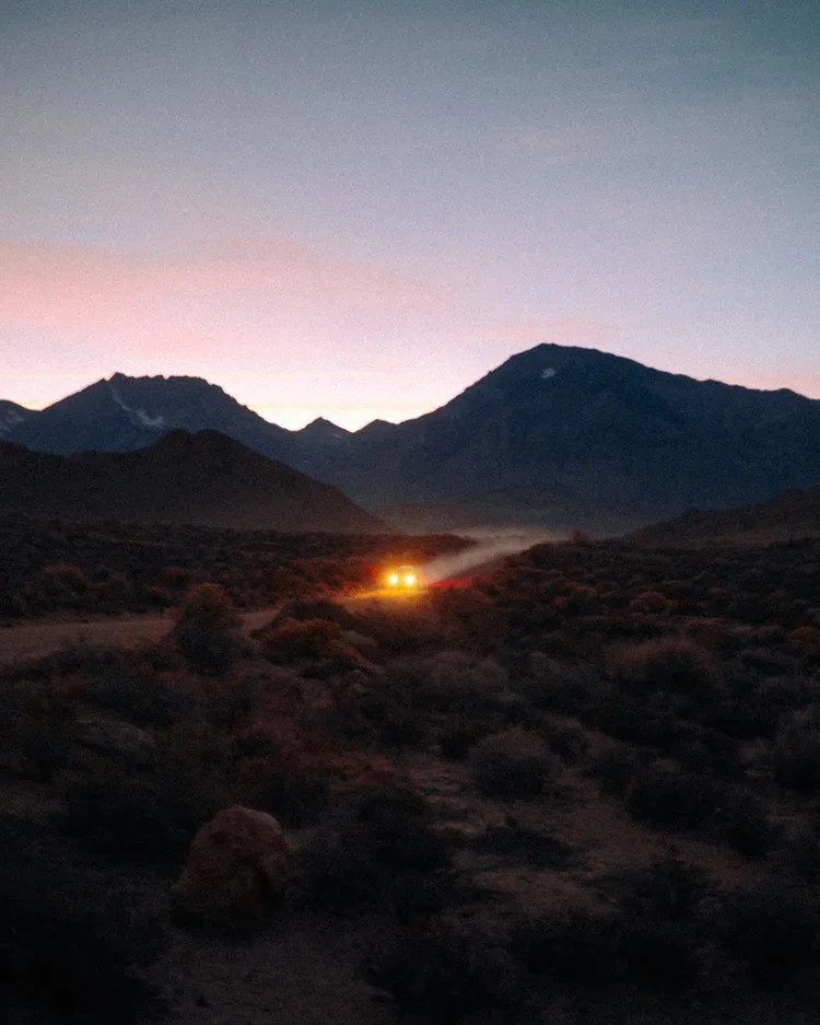 Dusk in a mountainous desert landscape with a sunset sky and a vehicle with headlights on, creating dust clouds.