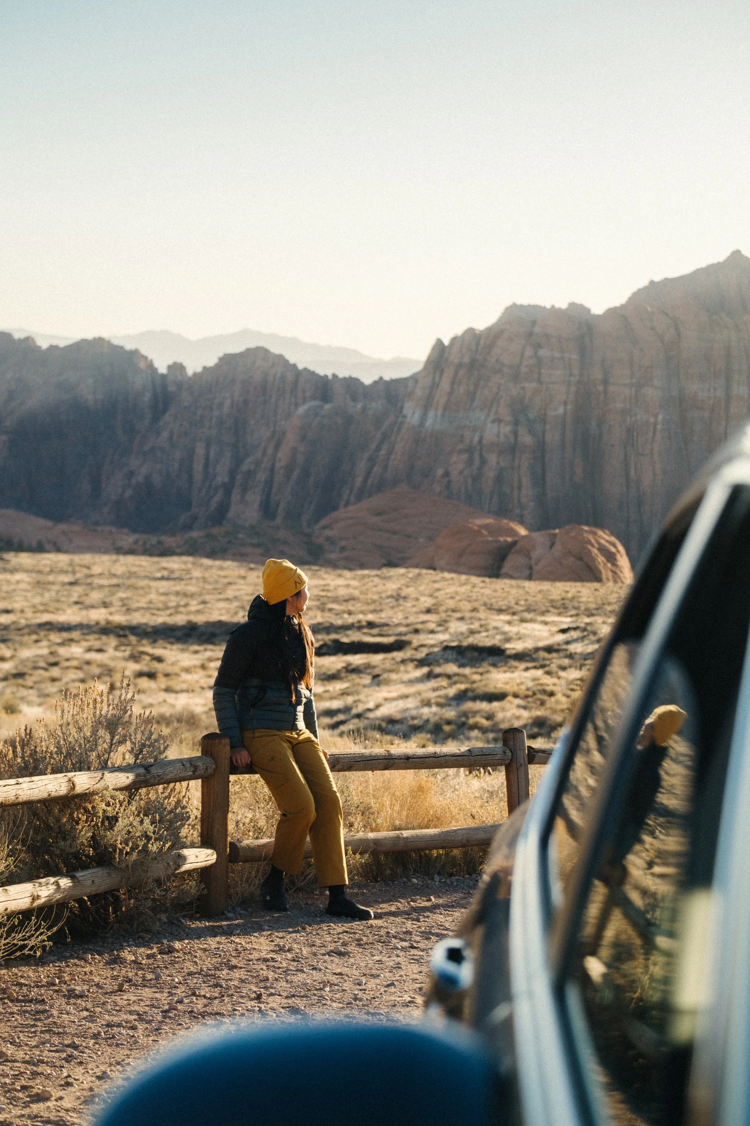 A woman sitting on a wooden fence in a desert landscape during sunset, with mountains in the background and a vehicle partially visible in the foreground.