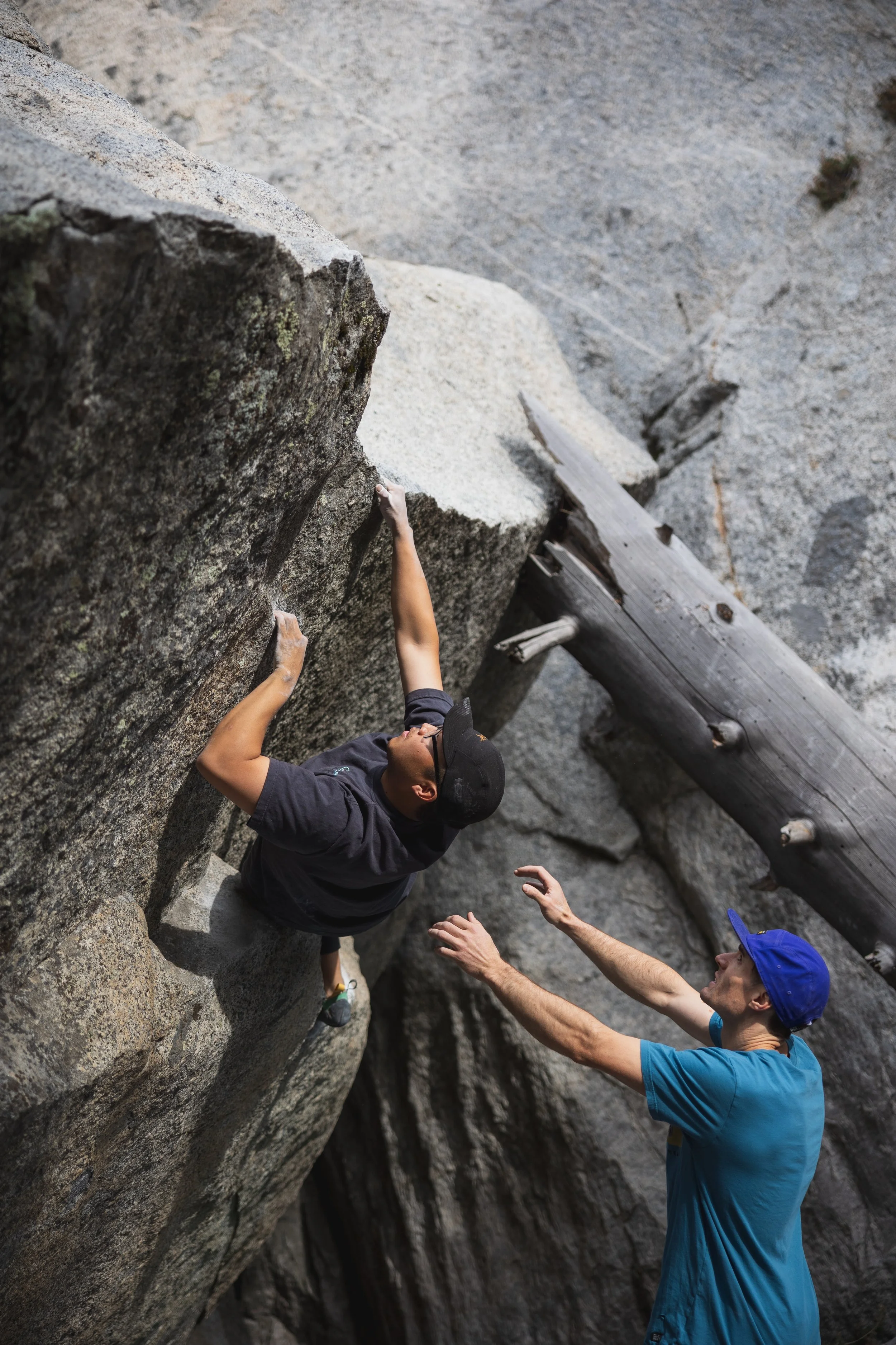 A man in a blue shirt and cap is spotting a woman rock climbing on a granite wall outdoors.
