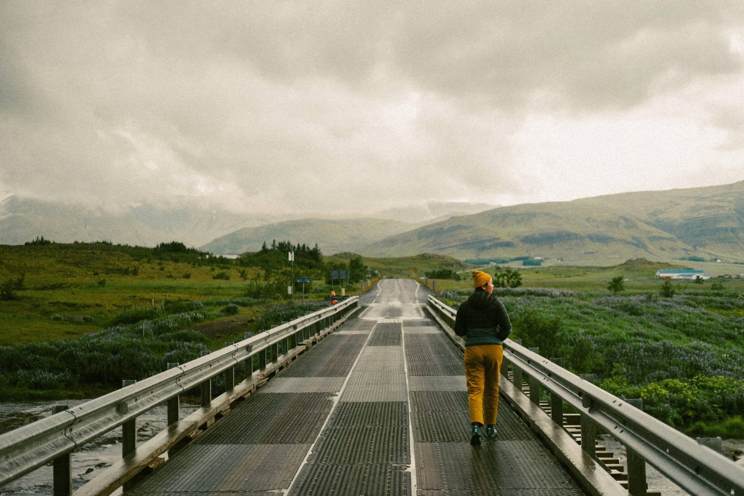 A person walking on a metal bridge in a rural landscape with green fields, hills, and mountains under cloudy sky.