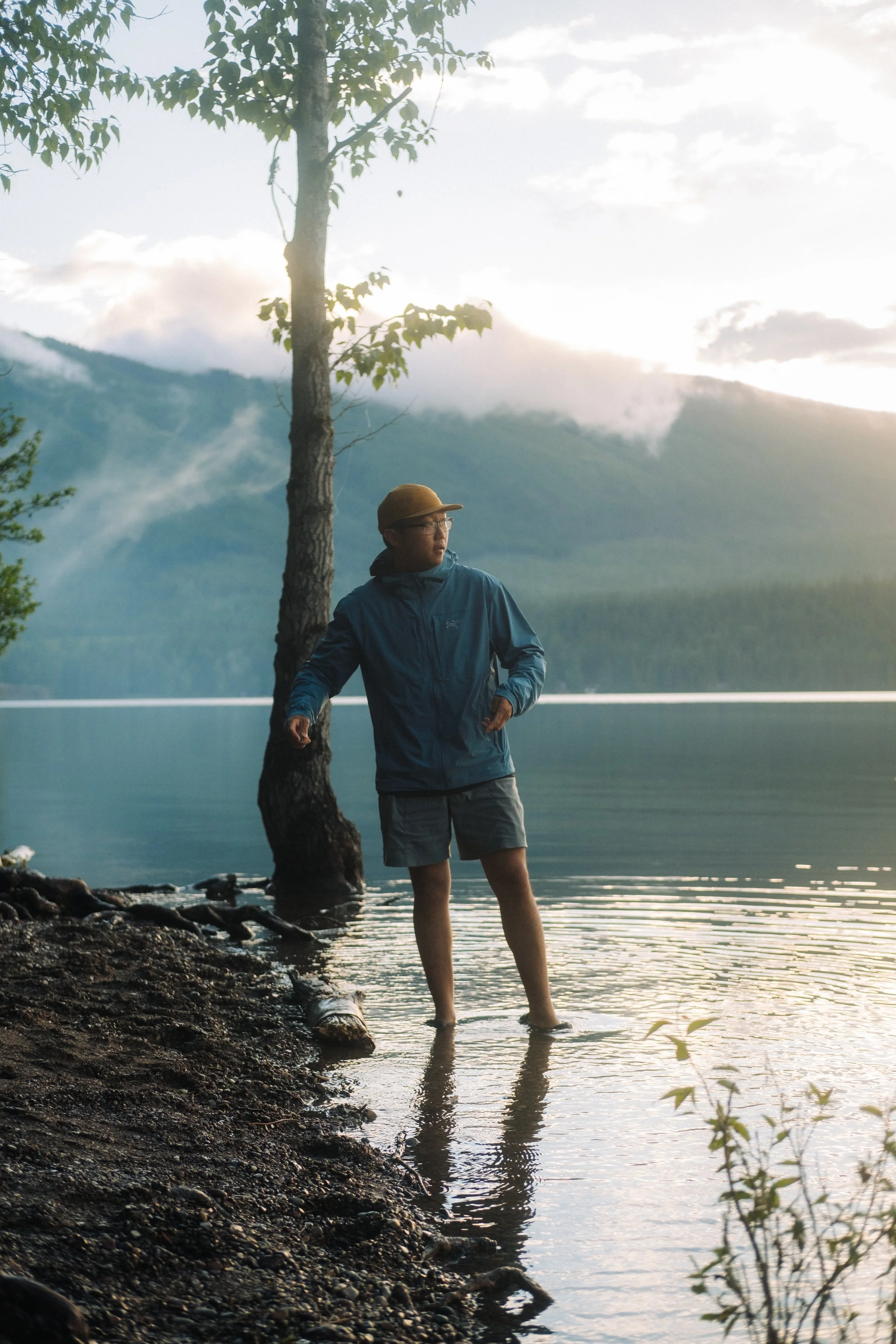 A person standing in the water near a lakeshore, surrounded by trees and mountains in the background, with the sky showing clouds at sunrise or sunset.