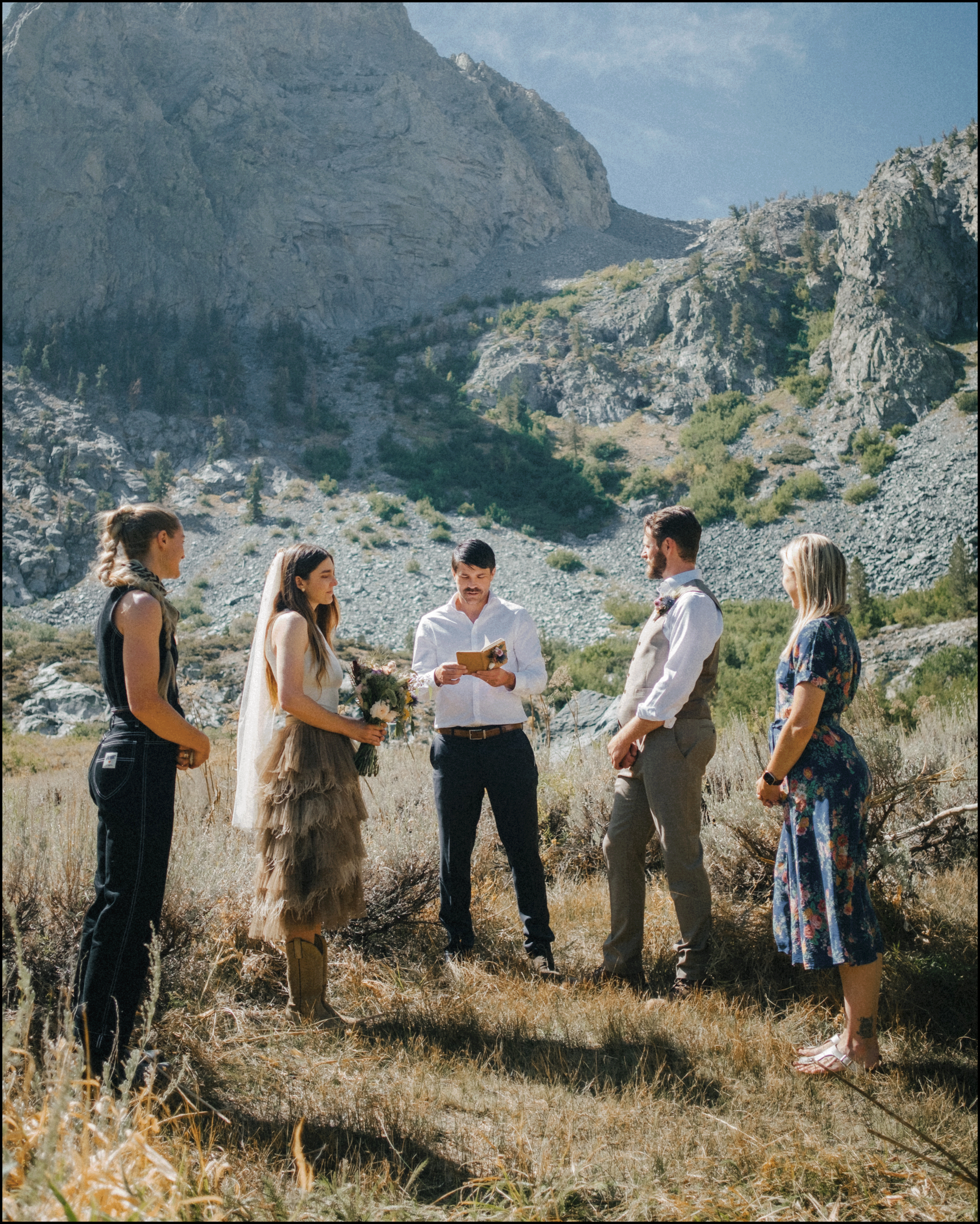 A wedding ceremony taking place outdoors in a mountainous area with six people standing on grass. The bride holds a bouquet and wears a tiered beige dress and veil. The officiant reads from a book, dressed in a white shirt. The groom is dressed in a light shirt and vest, and stands opposite the bride. Two women and one man stand on either side, dressed in casual or semi-formal attire. The backdrop features rocky mountains and blue sky.