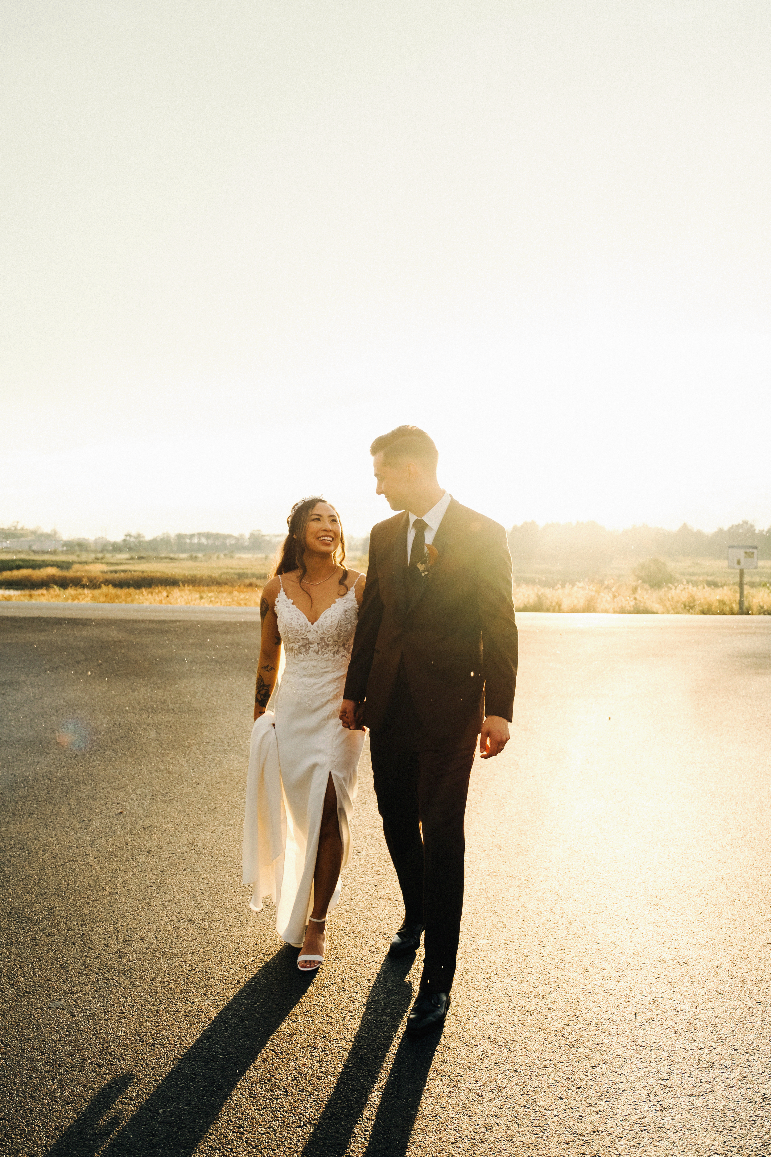 A newly married couple walking hand in hand on an empty road during sunset, with the bride in a white lace wedding dress and the groom in a black suit, smiling at each other.