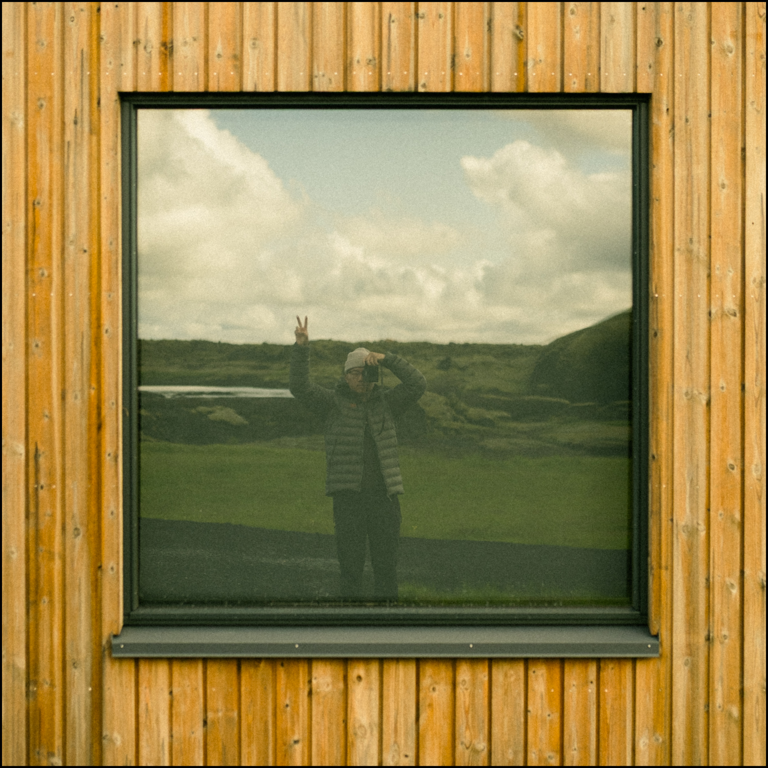 Reflection of a person in a window, holding a camera and making a peace sign, with a landscape of green hills and a cloudy sky in the background.