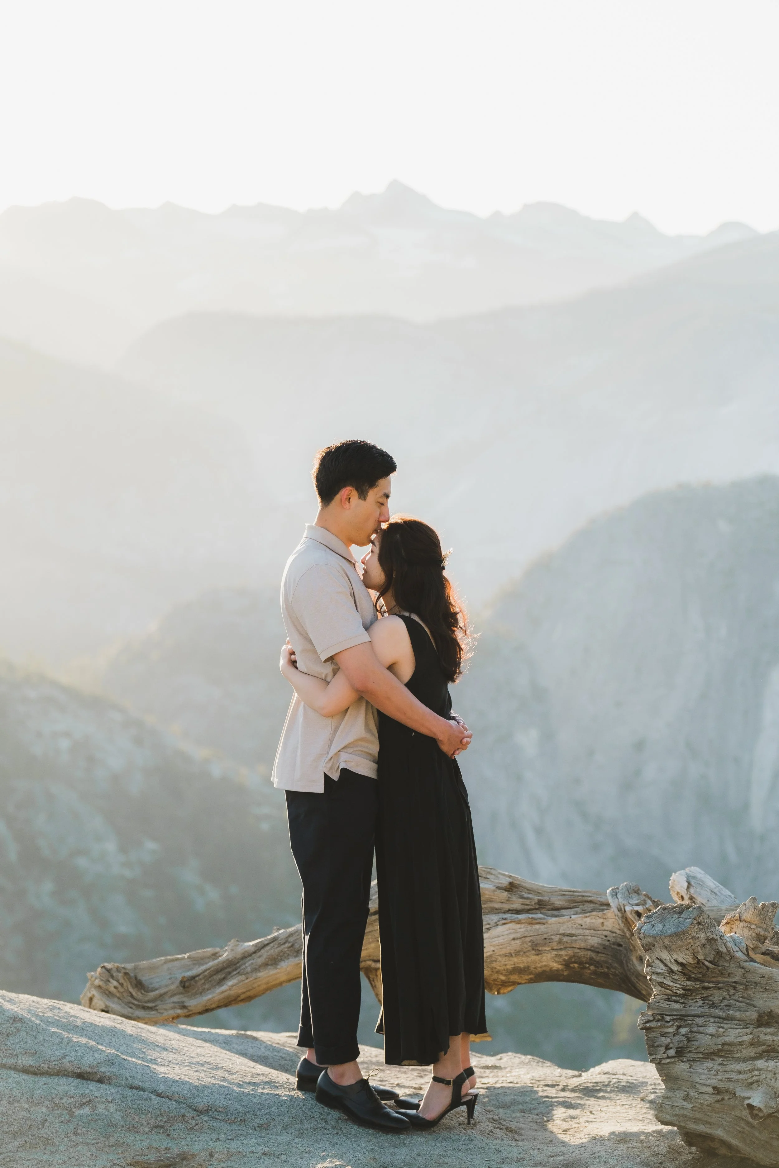 A couple embracing on a rocky ledge with mountains in the background during sunset.