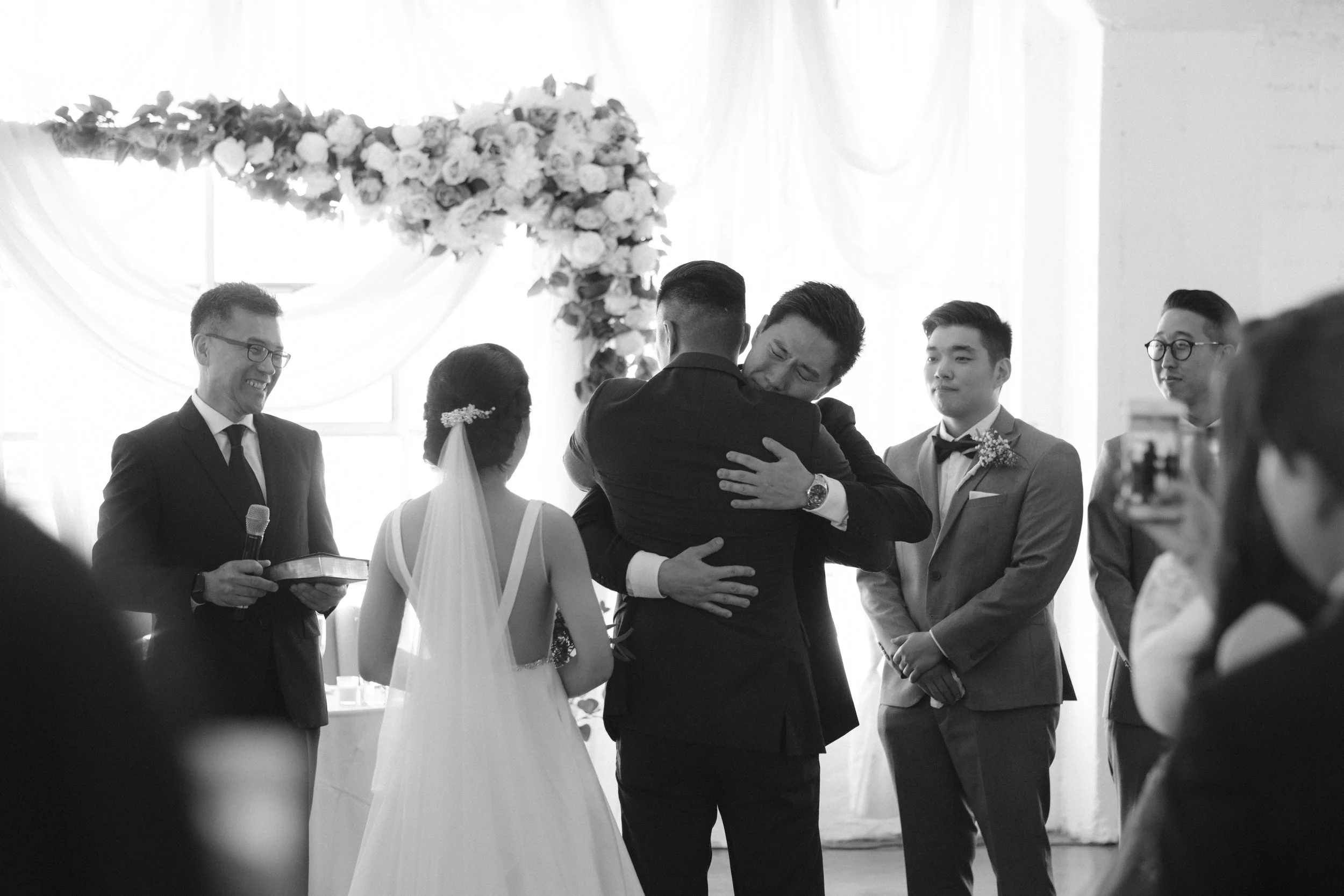 Groom and best man hugging during wedding ceremony with officiant and groomsmen watching.