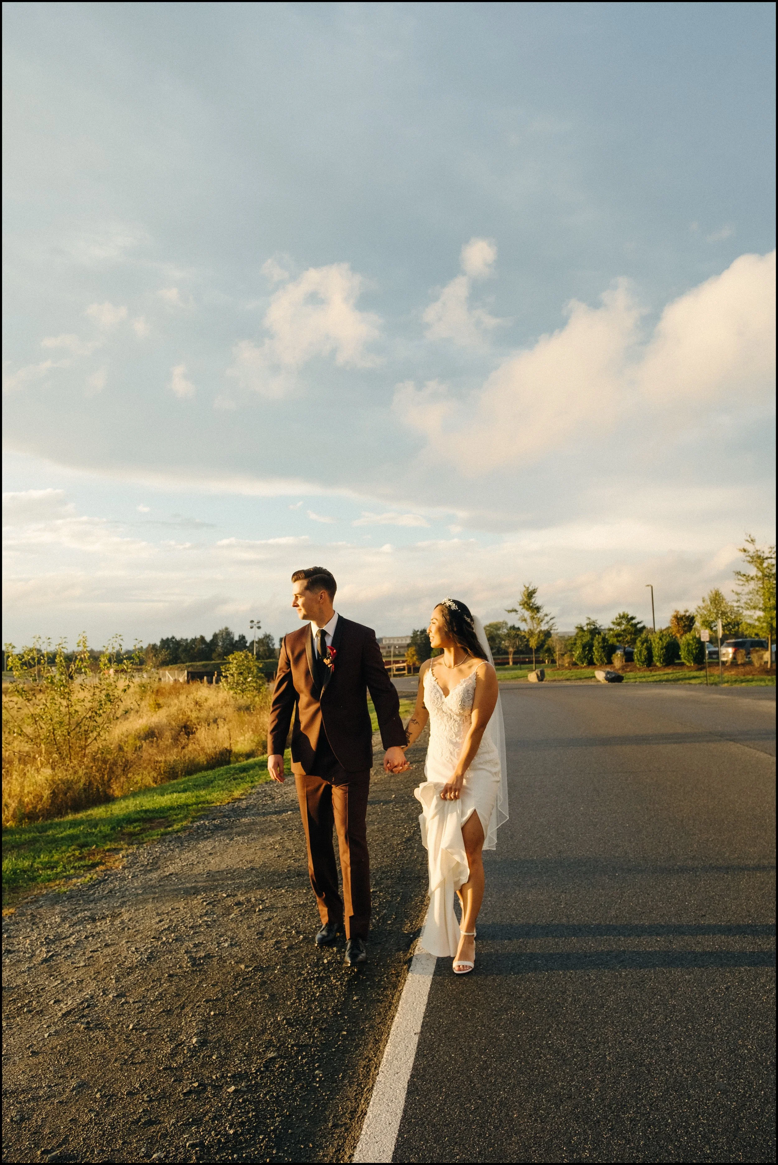 Bride and groom walking hand-in-hand on road during late afternoon, with green trees and a partly cloudy sky in the background.