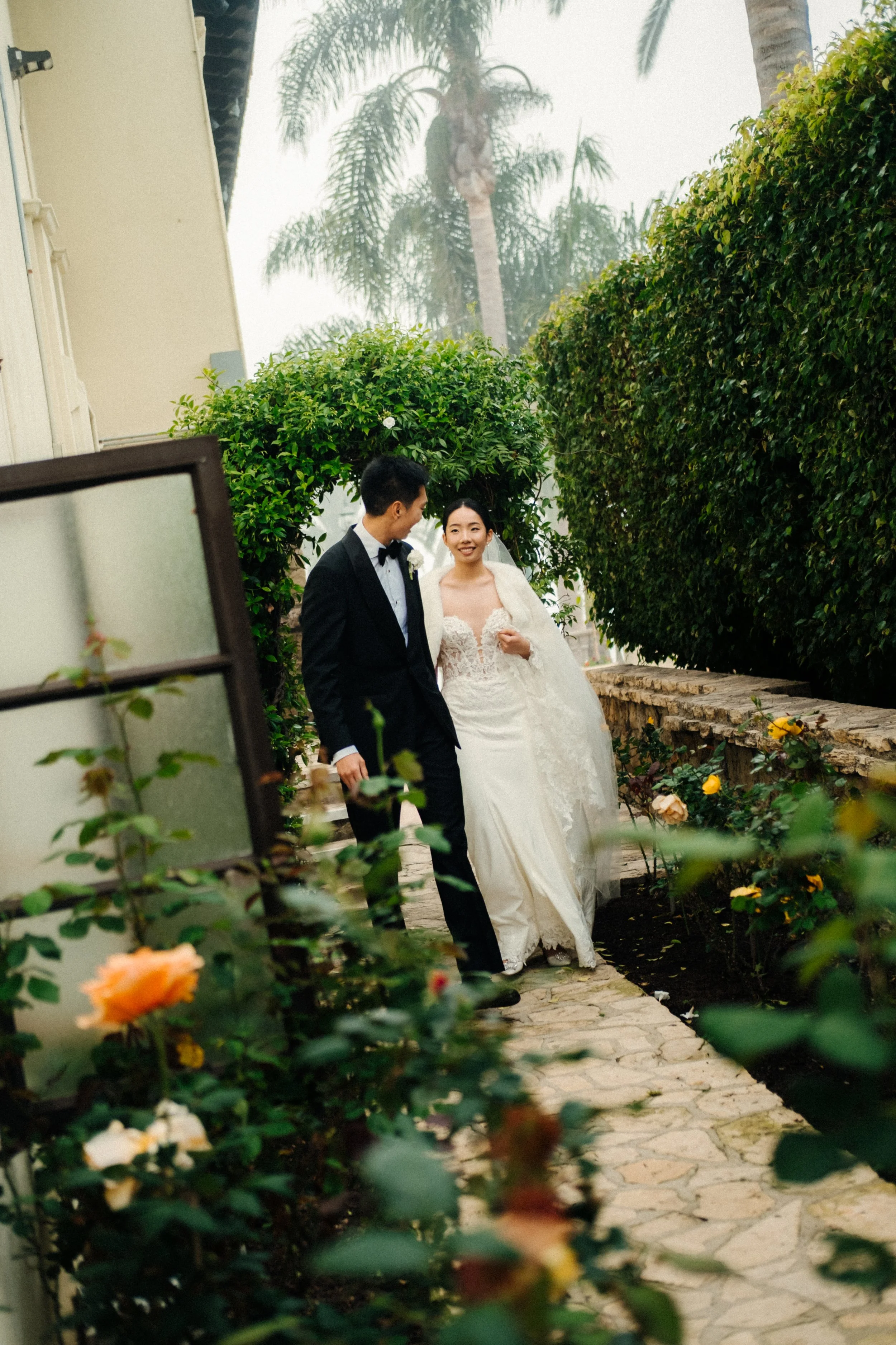 A bride and groom walking together outdoors in a garden, surrounded by greenery and flowers, with the bride in a white wedding gown and veil, and the groom in a black tuxedo and bow tie, enjoying a moment together.