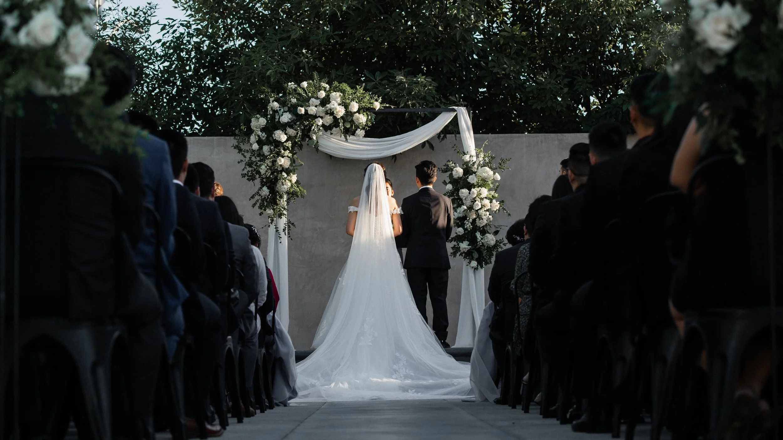 Bride and groom standing before an officiant at an outdoor wedding ceremony, with guests seated on either side and white floral decorations overhead.