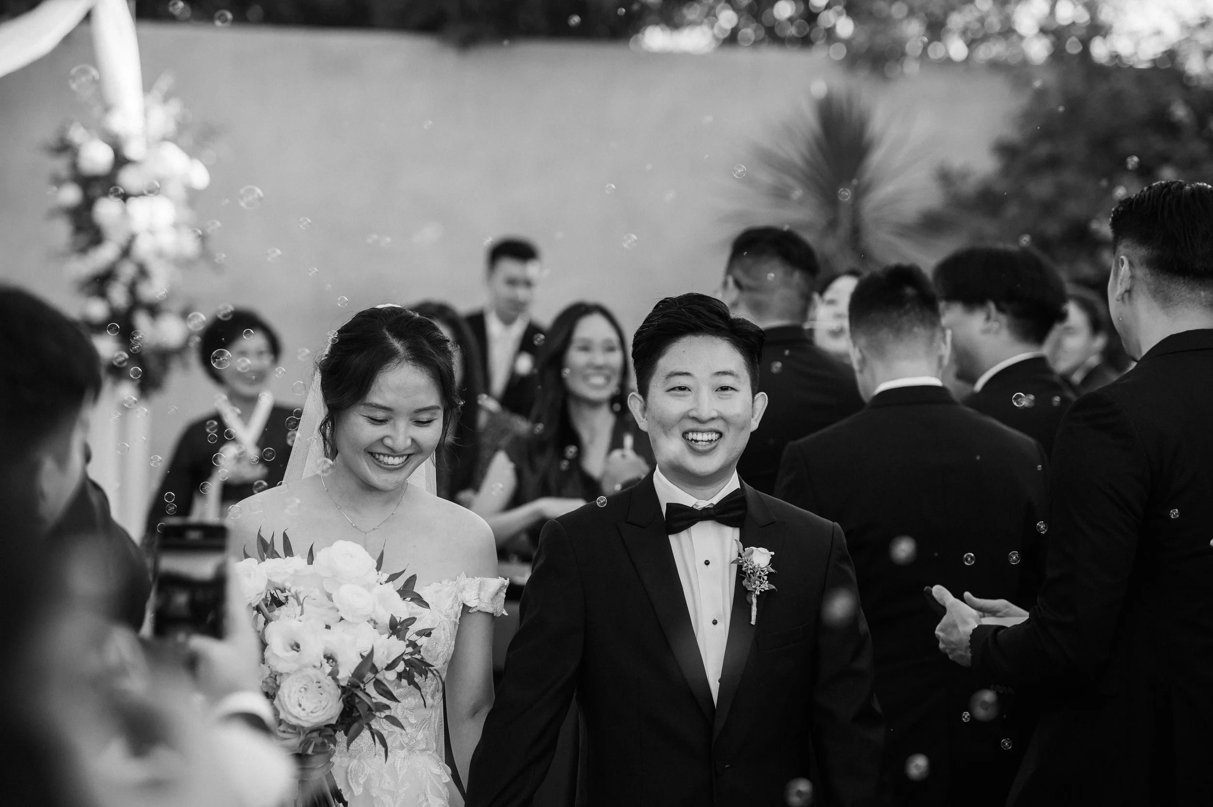 Black and white photo of a wedding celebration with a smiling bride and groom in the center, surrounded by guests, some holding phones, with bubbles floating in the air.