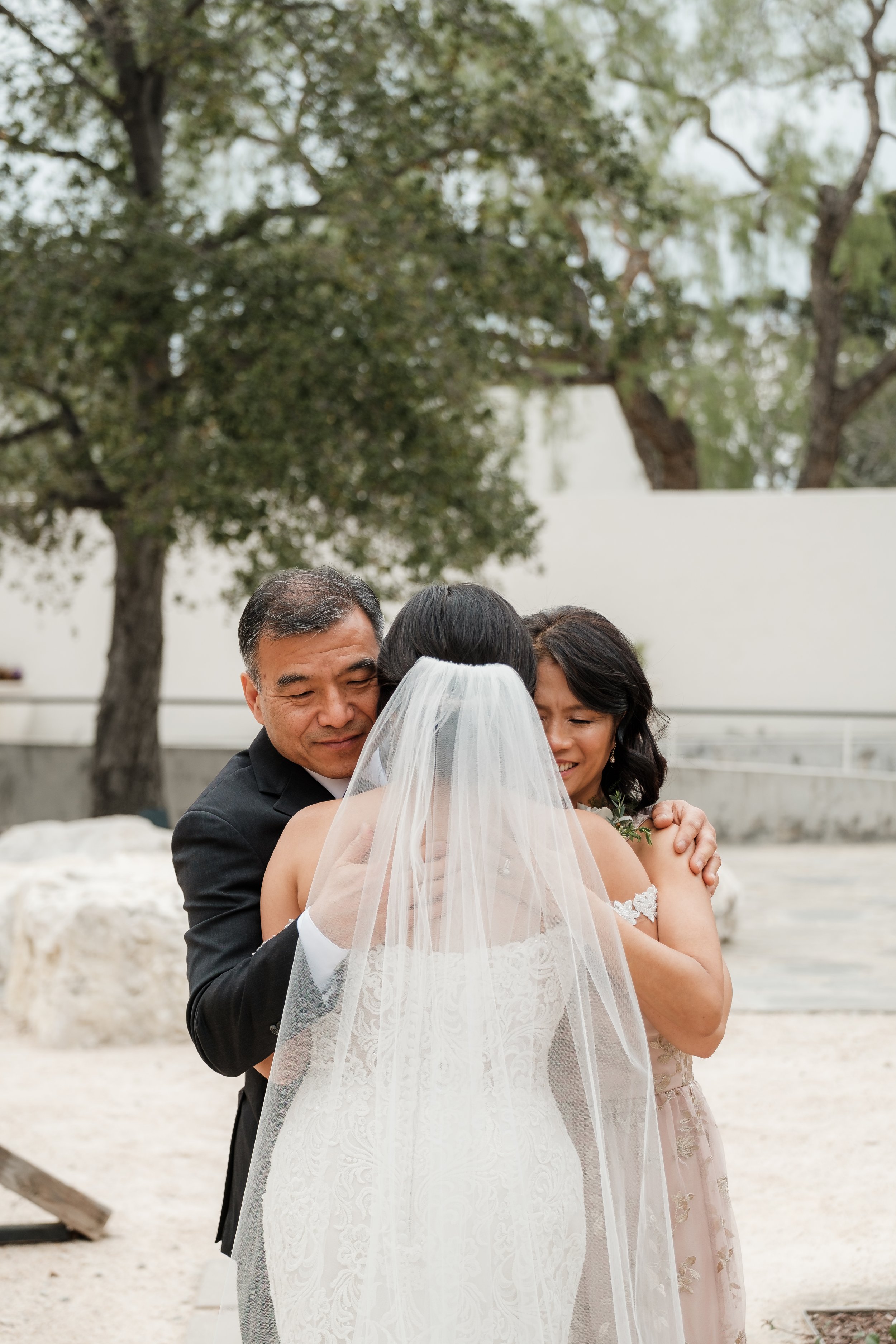 A bride, dressed in a wedding gown and veil, hugs her parents outdoors during her wedding moment.